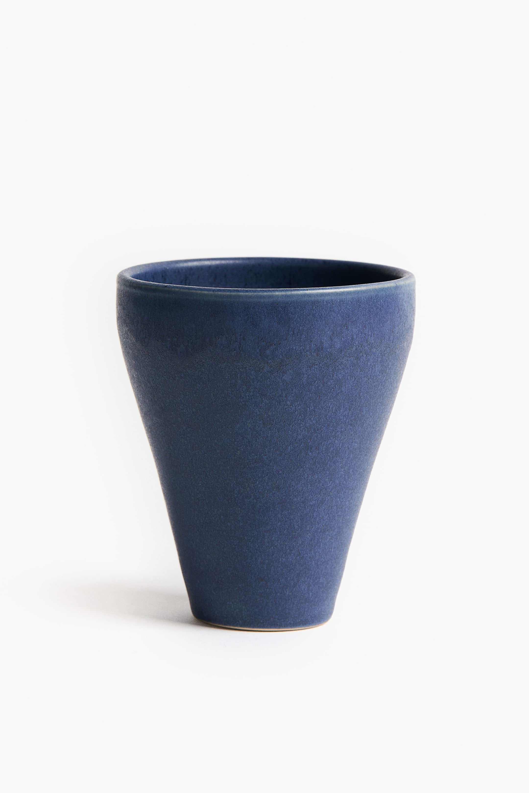 Zobacz większy obraz: Niebieski ceramiczny kubek stoi pionowo, ukazując zwężający się, stożkowy kształt, który zwęża się ku płaskiej podstawie. Kubek ma teksturowane matowe szkliwo w głębokim indygo, z cienkim pierścieniem nieglazurowanej beżowej gliny widocznym na samym dole.