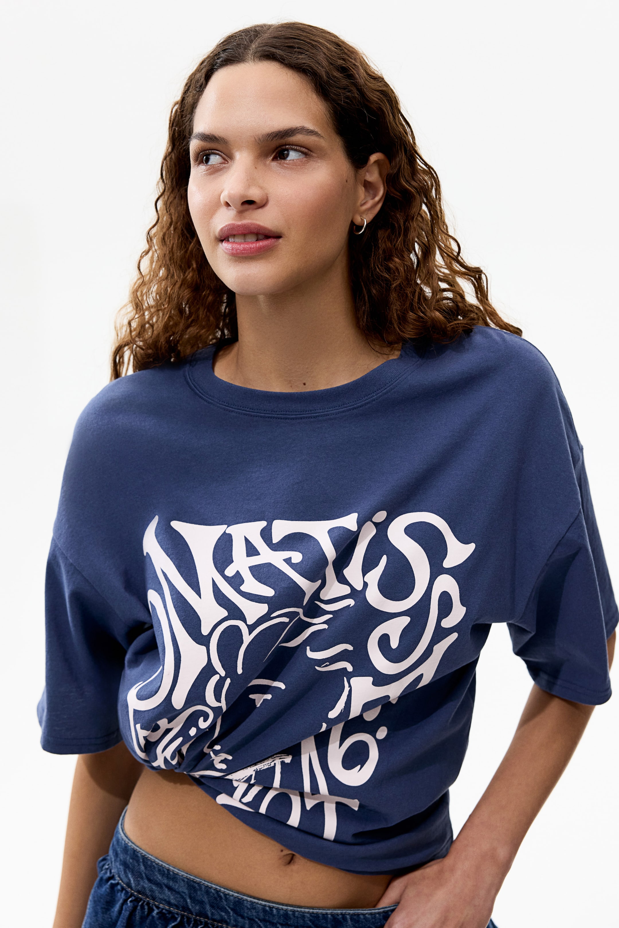 View larger image: A blue crew neck t-shirt menampilkan reka bentuk grafik putih yang besar di bahagian hadapan. T-shirt tersebut diikat simpul di bahagian hem depan, menghasilkan potongan cropped.