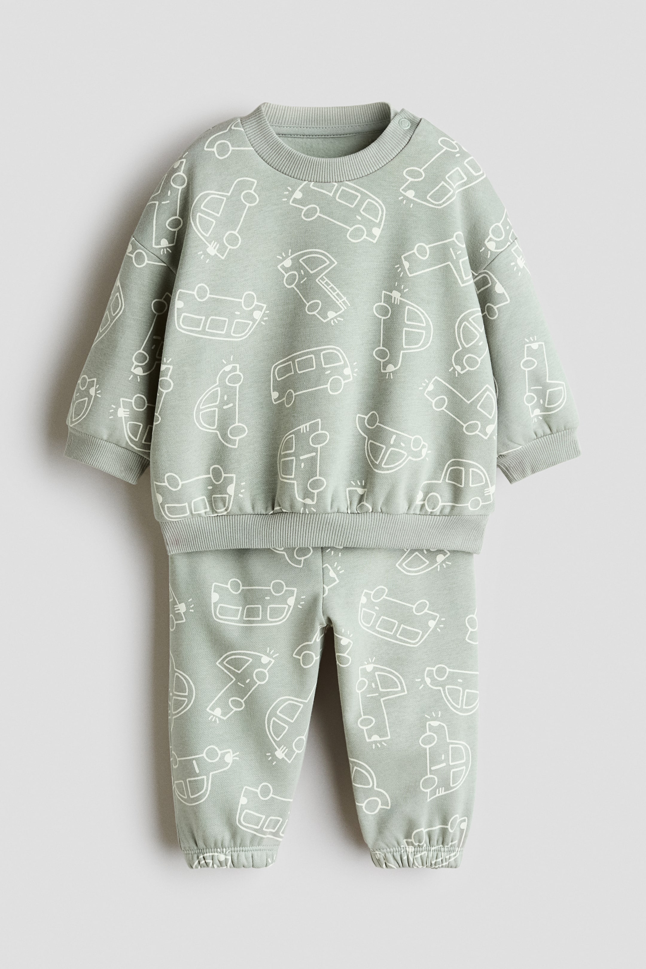 View larger image: The photo showcases a 2-piece printed sweatshirt set from the front, highlighting a light green hue adorned with white vehicle outlines. The sweatshirt features a ribbed crew neck, ribbed cuffs, a ribbed hem, and snap buttons on one shoulder. The coordinating pants come with ribbed ankle cuffs.