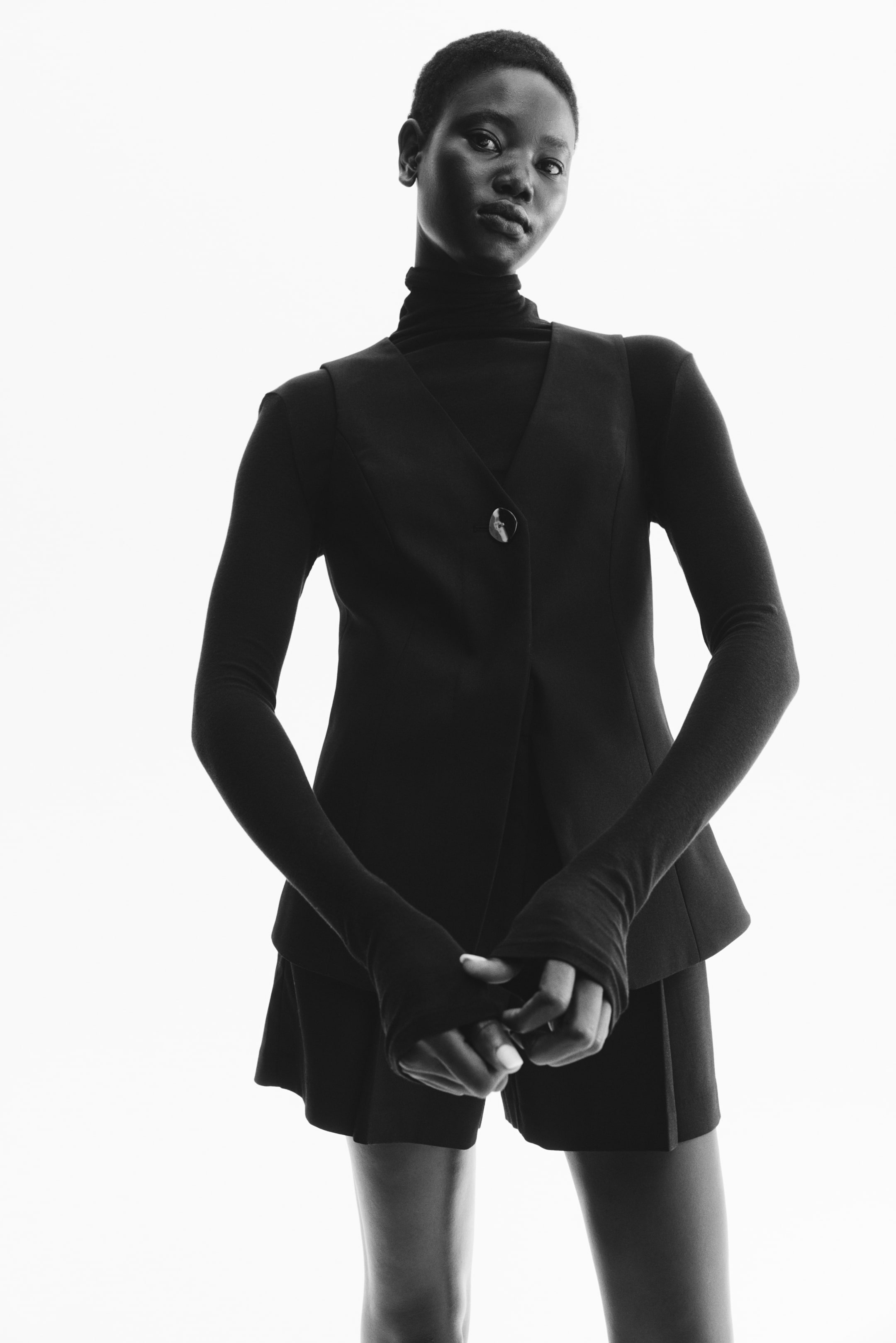 Ampliar la imagen: Se lleva puesto un chaleco negro sin mangas orientado hacia el frente, con un escote en V y un solo botón de cierre claro y descentrado, sobre un suéter de cuello alto negro de manga larga con mangas extendidas que cubren las manos, combinado con pantalones cortos negros entallados visibles por debajo del dobladillo del chaleco.