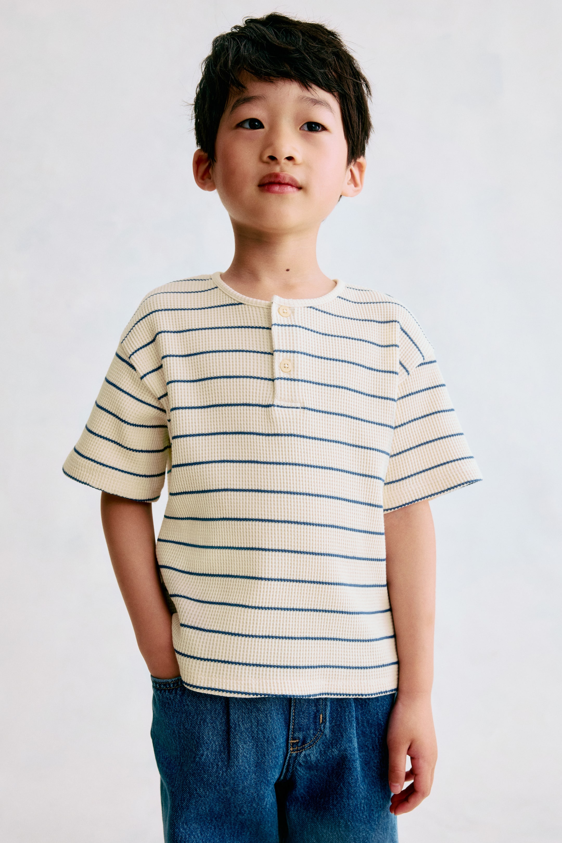 Ver imagen más grande: Se muestra desde el frente un top Henley de jersey en color crema con textura de gofre y rayas horizontales en azul marino. Presenta un escote redondo con un ribete estrecho, una tapeta con dos botones, hombros caídos y mangas cortas.