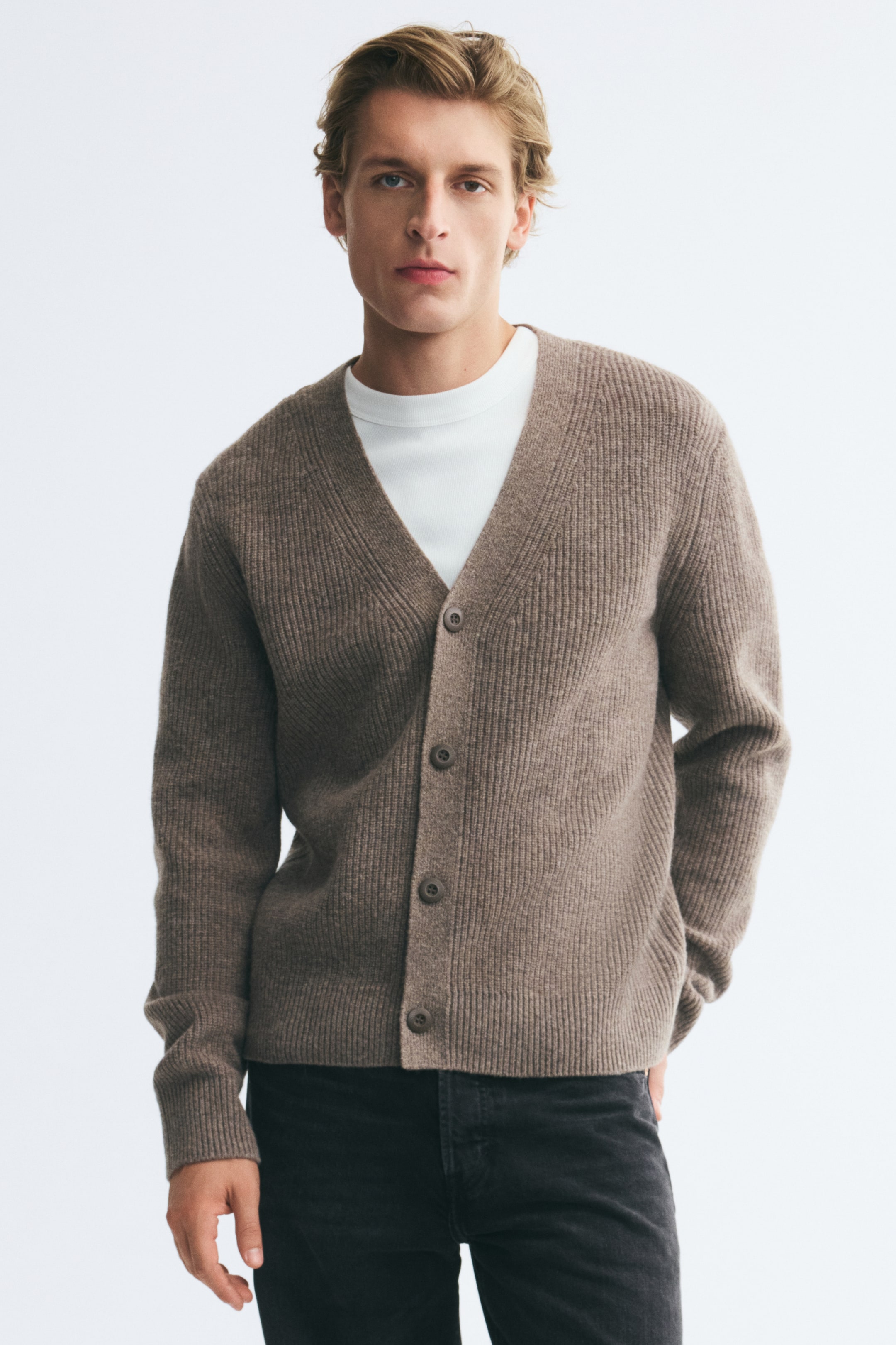 Vezi imaginea mai mare: Un cardigan din lână, cu tricotaj rib, de culoare maro, este purtat de model peste un tricou alb cu guler rotund și blugi gri închis. Cardiganul are un decolteu în V, închidere cu nasturi în față și mâneci lungi cu manșete striate.