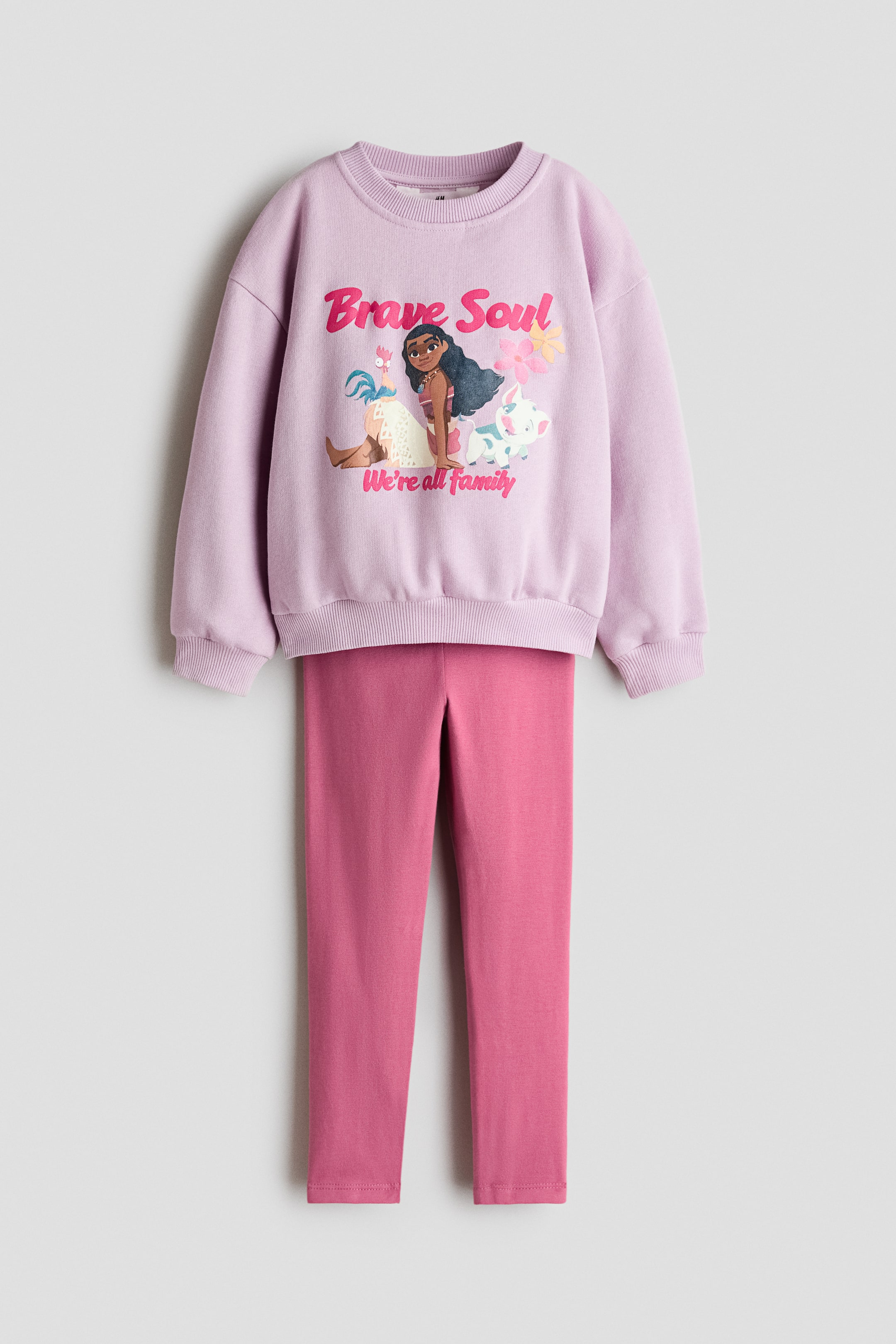 Ver imagem maior: Vê-se um conjunto de duas peças pela frente, composto por uma sweatshirt lilás clara e leggings rosa-escuro coordenadas. A sweatshirt apresenta uma estampagem gráfica de Moana e os seus companheiros animais no peito e foi concebida com gola redonda canelada, ombros descaídos e punhos e bainha canelados.