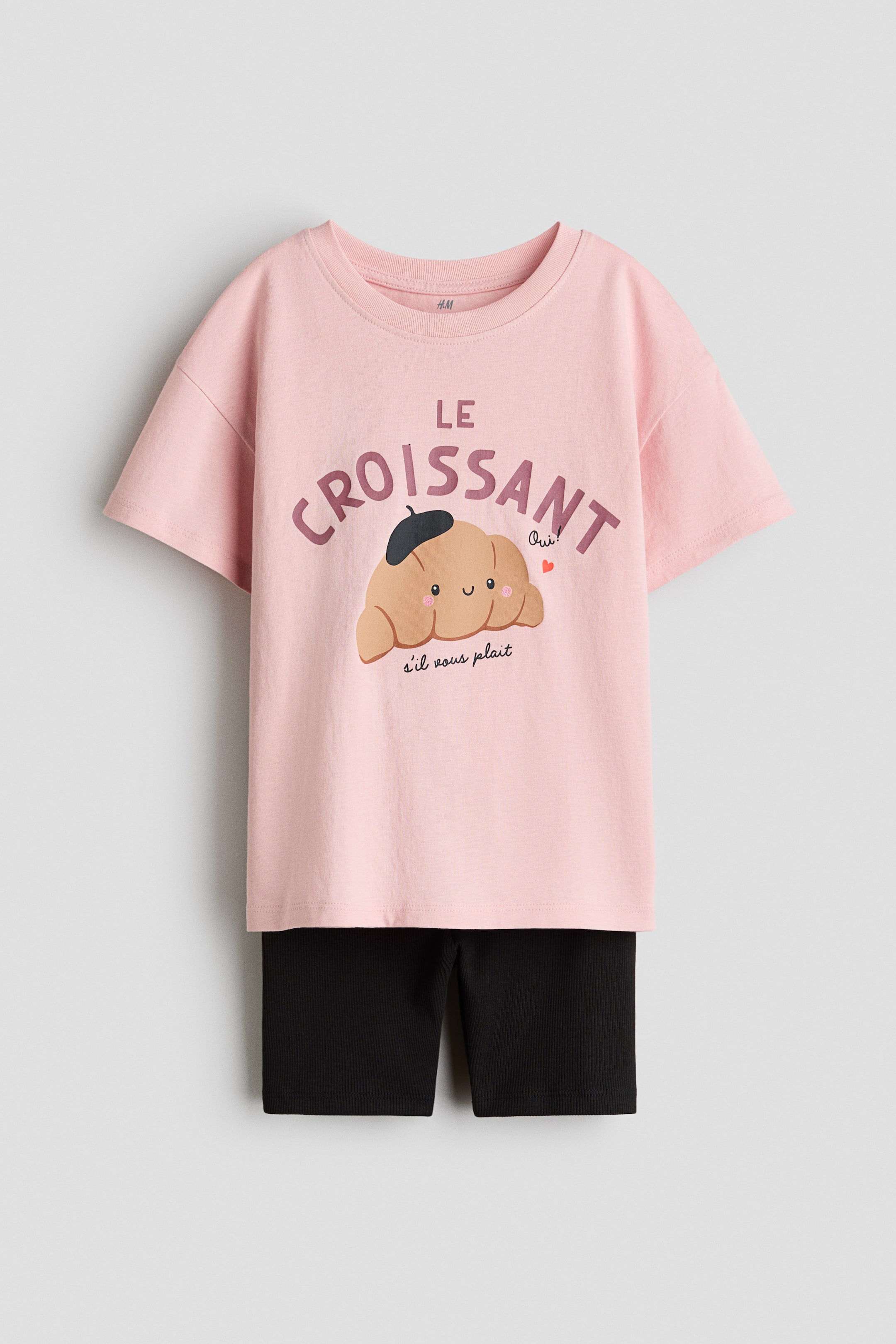 Größeres Bild ansehen: Ein Set aus einem rosa T-Shirt und schwarzen Radlerhosen wird von vorne präsentiert. Das T-Shirt zeigt einen "LE CROISSANT"-Grafikdruck, einen gerippten Halsausschnitt und überschnittene Schultern, während die schwarzen Radlerhosen eine gerippte Textur und einen verdeckten elastischen Bund aufweisen.
