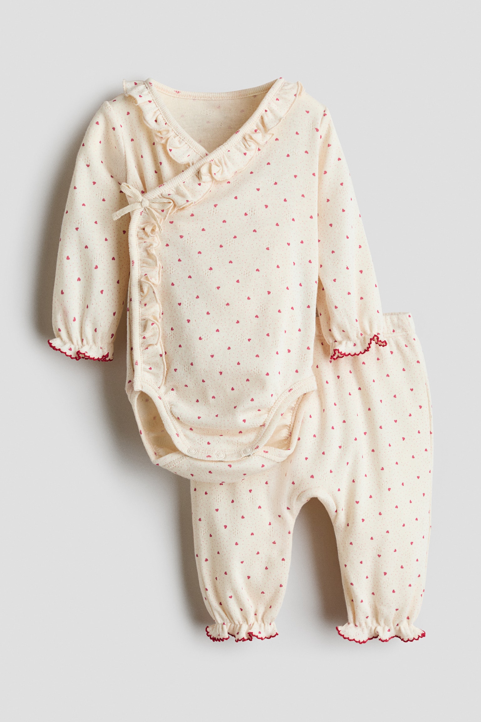Un ensemble de vÃªtements pour bÃ©bÃ© est prÃ©sentÃ© de face, comprenant un body croisÃ© de couleur crÃ¨me et un pantalon assorti imprimÃ© de minuscules cÅurs rouges. Le body Ã  manches longues prÃ©sente des finitions volantÃ©es avec une bordure picot rouge, un nÅud dÃ©coratif sur le cÃ´tÃ© et des boutons-pression Ã  lâentrejambe, tandis que le pantalon assorti montre des ourlets Ã©lastiquÃ©s et volantÃ©s.