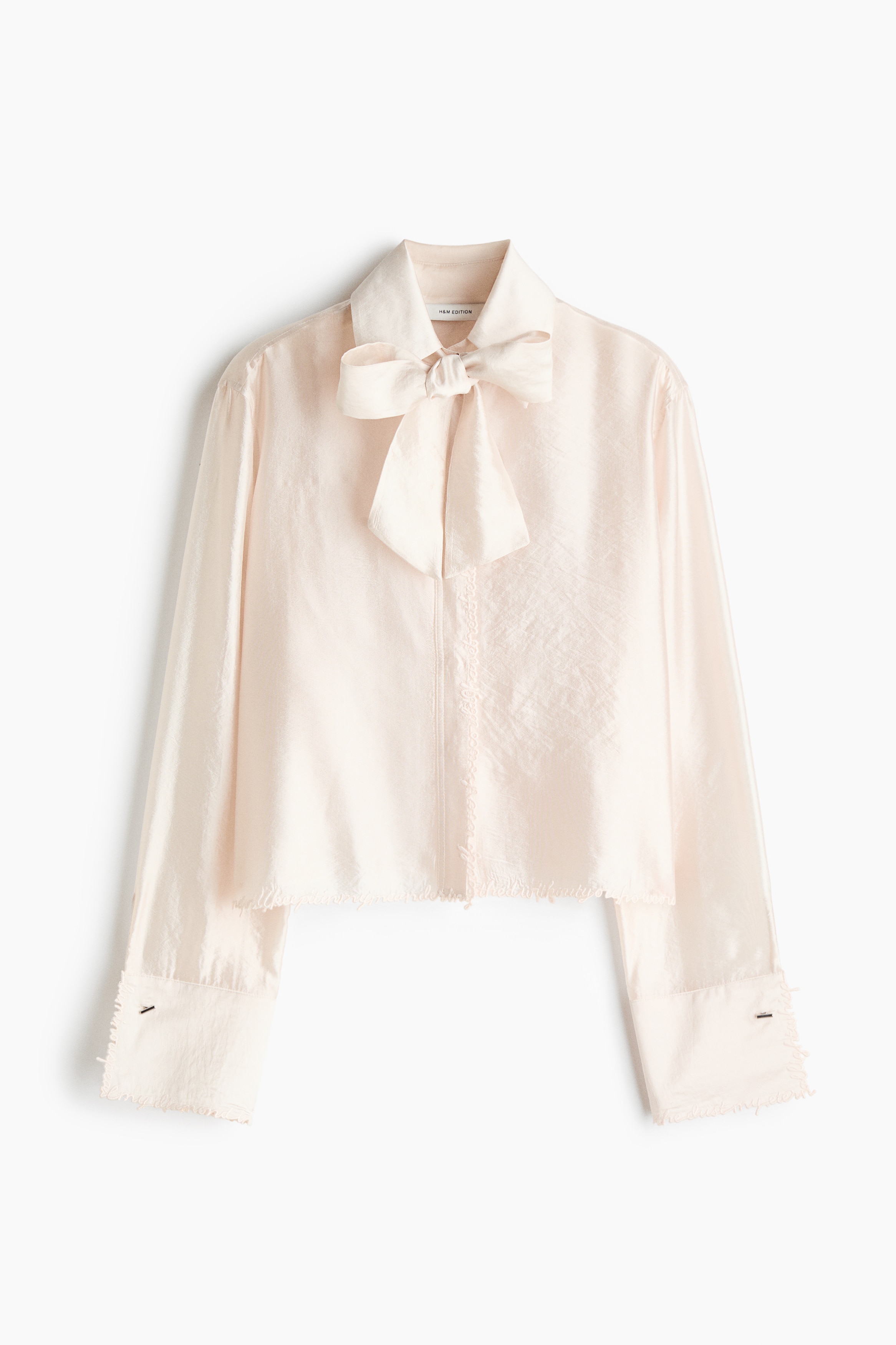 Une chemise semi-transparente blanc cassÃ©, lÃ©gÃ¨rement satinÃ©e, est portÃ©e par le mannequin, associÃ©e Ã  un pantalon noir. La chemise prÃ©sente un col montant avec un nÅud drapÃ©, des manches longues avec de larges poignets effilochÃ©s, ainsi que des dÃ©tails de texte brodÃ©s le long de lâourlet infÃ©rieur.