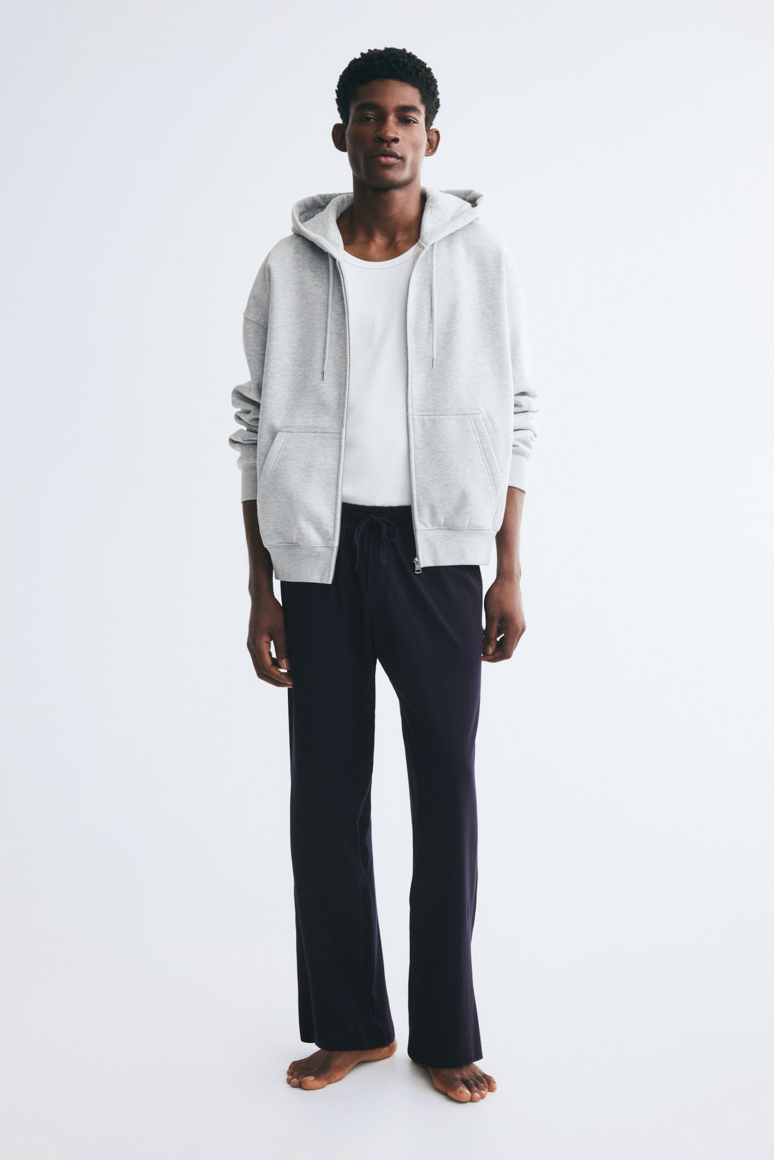 The model is wearing dark-coloured cotton jersey trousers featuring a relaxed, straight-leg fit and an elasticated drawstring waist. The trousers are styled with a tucked-in white T-shirt and an open light grey zip-up hoodie. - 1