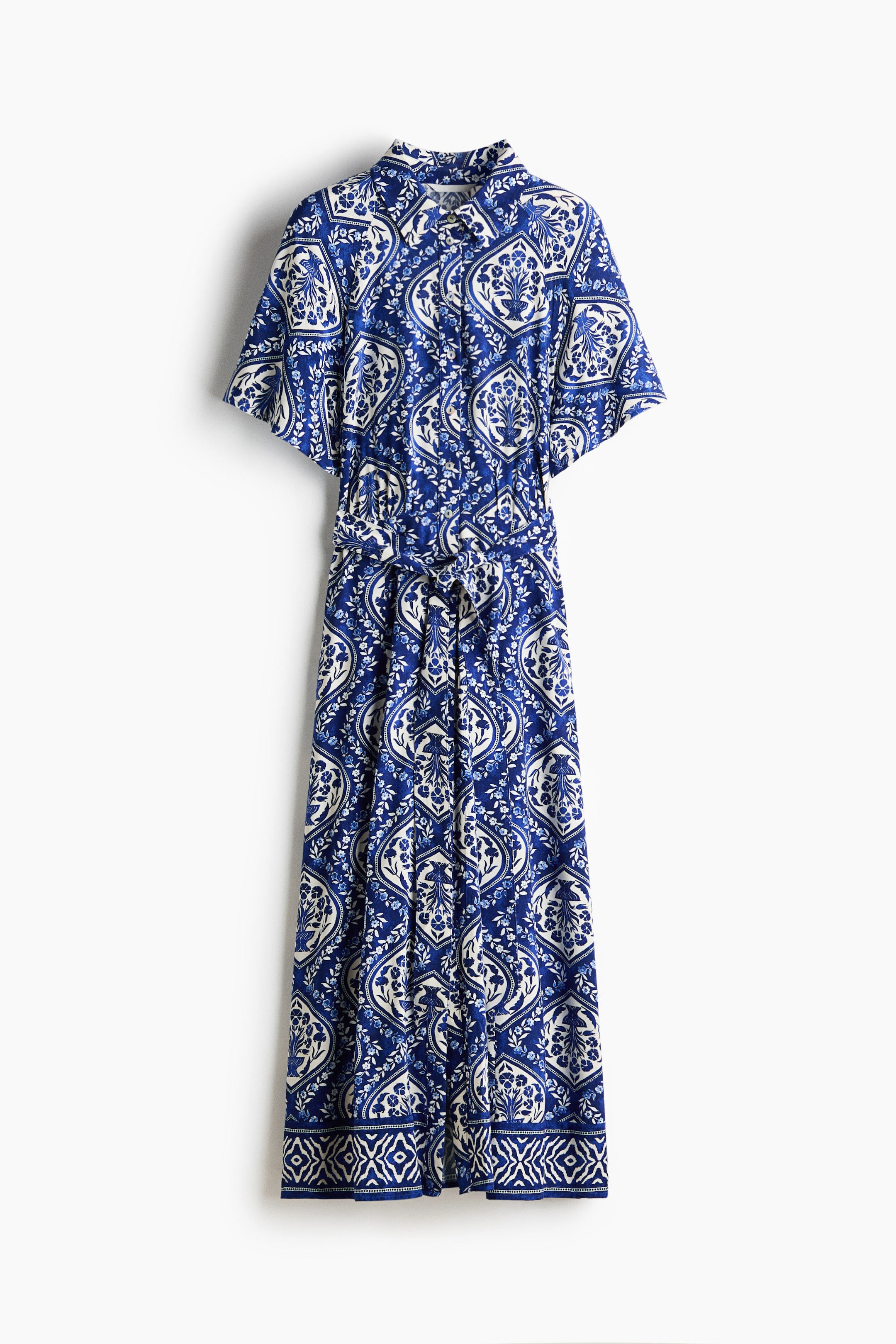 View larger image: The front of a calf-length dress showcases an intricate blue and white pattern, a collar, and buttons running down the front. It features short raglan sleeves, gathers at the front waist, and a detachable tie belt knotted at the center.