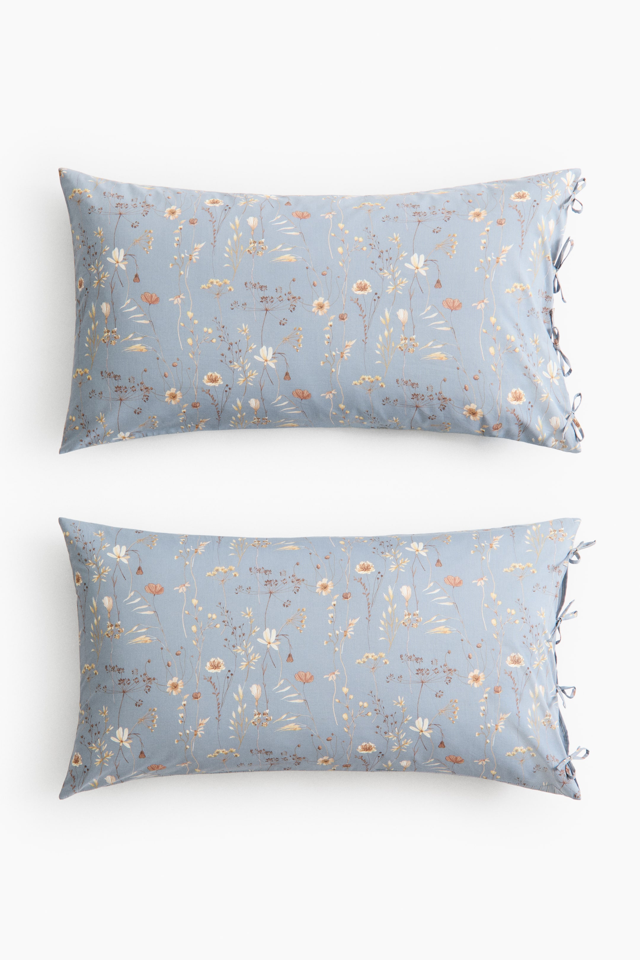Ver imagen más grande: Dos fundas de almohada muestran un estampado floral en su parte delantera, con pequeñas flores y follaje en tonos blanco, crema, amarillo y naranja claro sobre un fondo azul claro. Se pueden ver múltiples lazos a lo largo del lado derecho de cada funda de almohada.