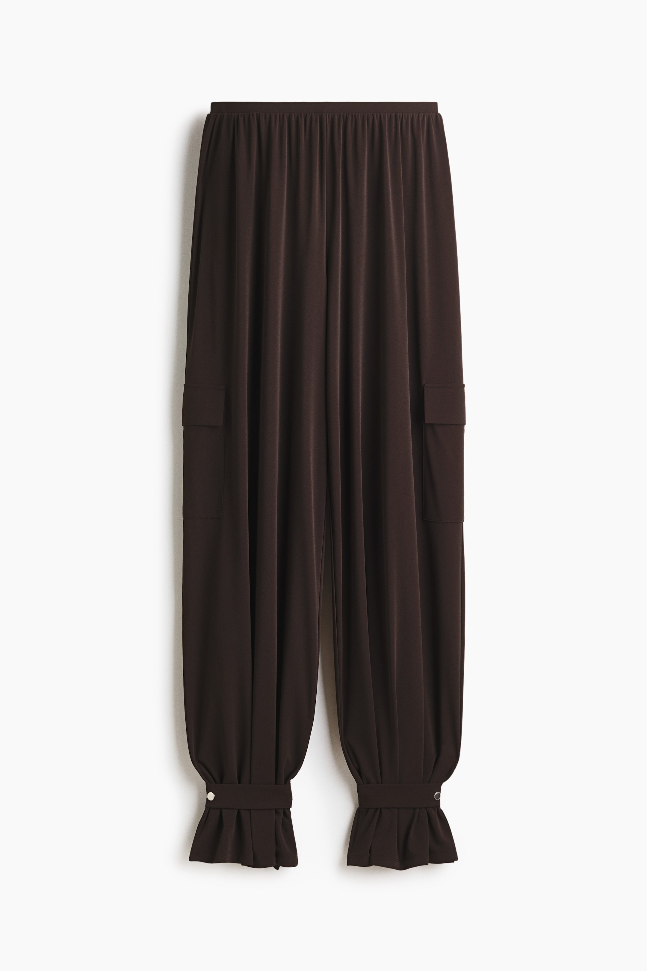 Visa större bild: Mörkbruna byxor med en rynkad elastisk midja, löst sittande ben med två fickor med lock, och rynkade muddar med dekorativa tryckknappar och volangkant.