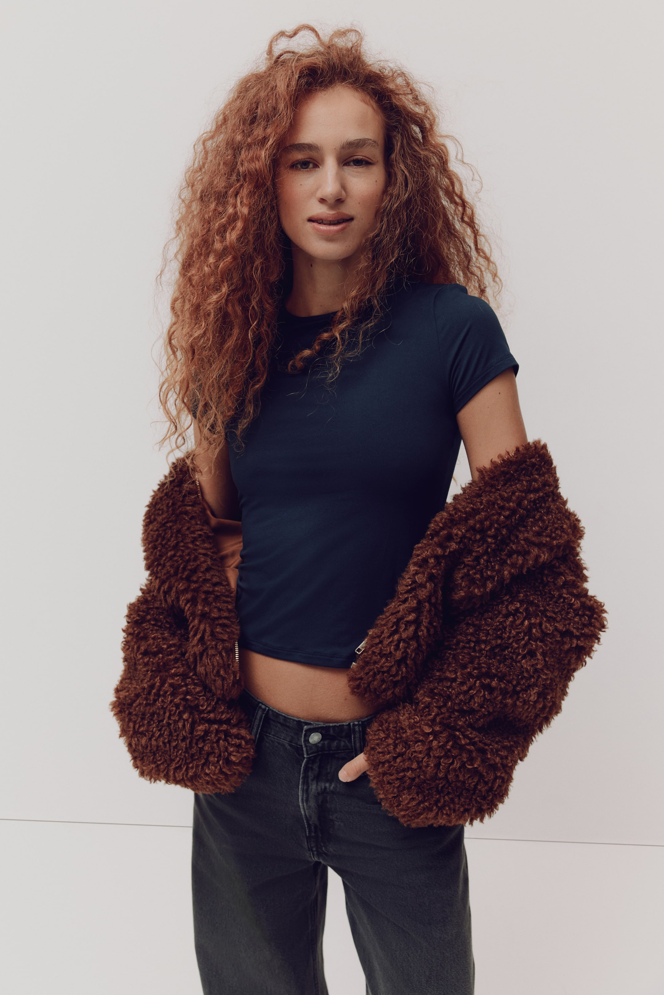 Voir une image plus grande: Un t-shirt à manches courtes bleu marine présente un col rond et une coupe courte et ajustée; il est présenté de face et visible sous une veste brune duveteuse posée sur les épaules.