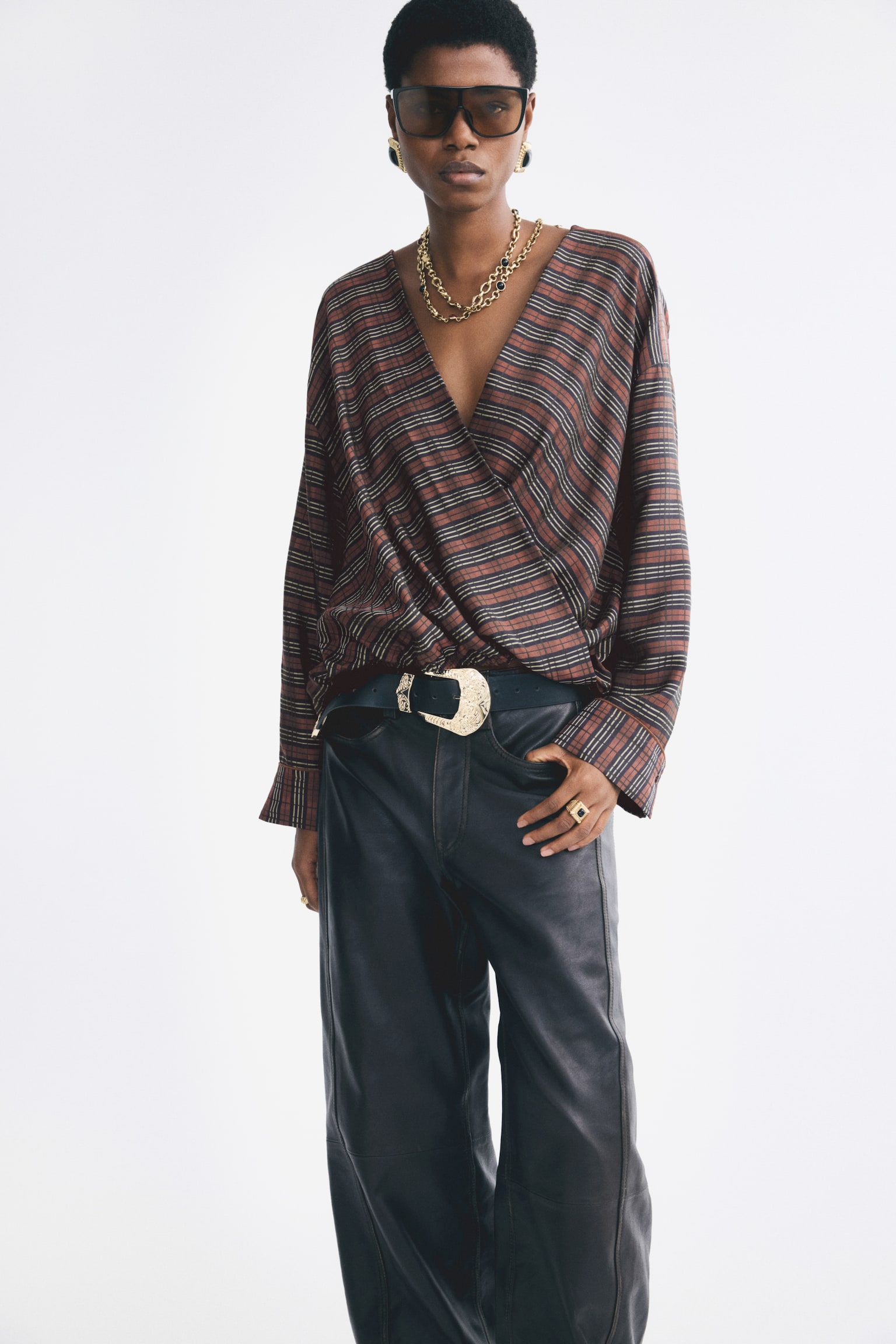 The model is wearing an oversized blouse with a brown and black striped pattern, featuring a deep V-neckline and a wrap-front design. The long-sleeved blouse is tucked into dark leather trousers, styled with a large-buckled belt. - 1