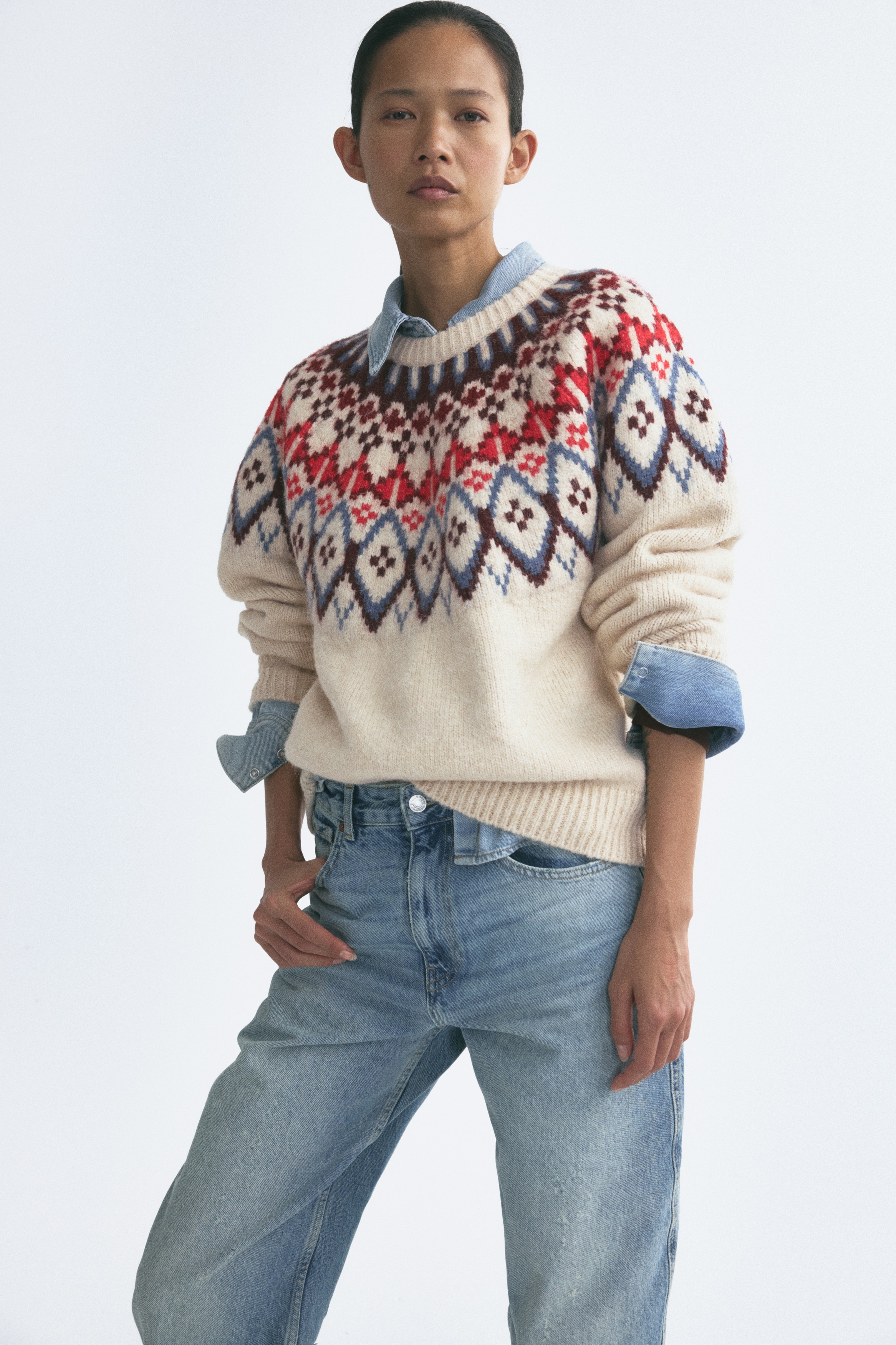 A cream-colored knit sweater is shown from the front, featuring a multi-colored Fair Isle yoke pattern in red, blue, and burgundy around the crew neck, shoulders, and upper sleeves. The sweater is finished with a ribbed neckline, ribbed cuffs, and a ribbed hem.
