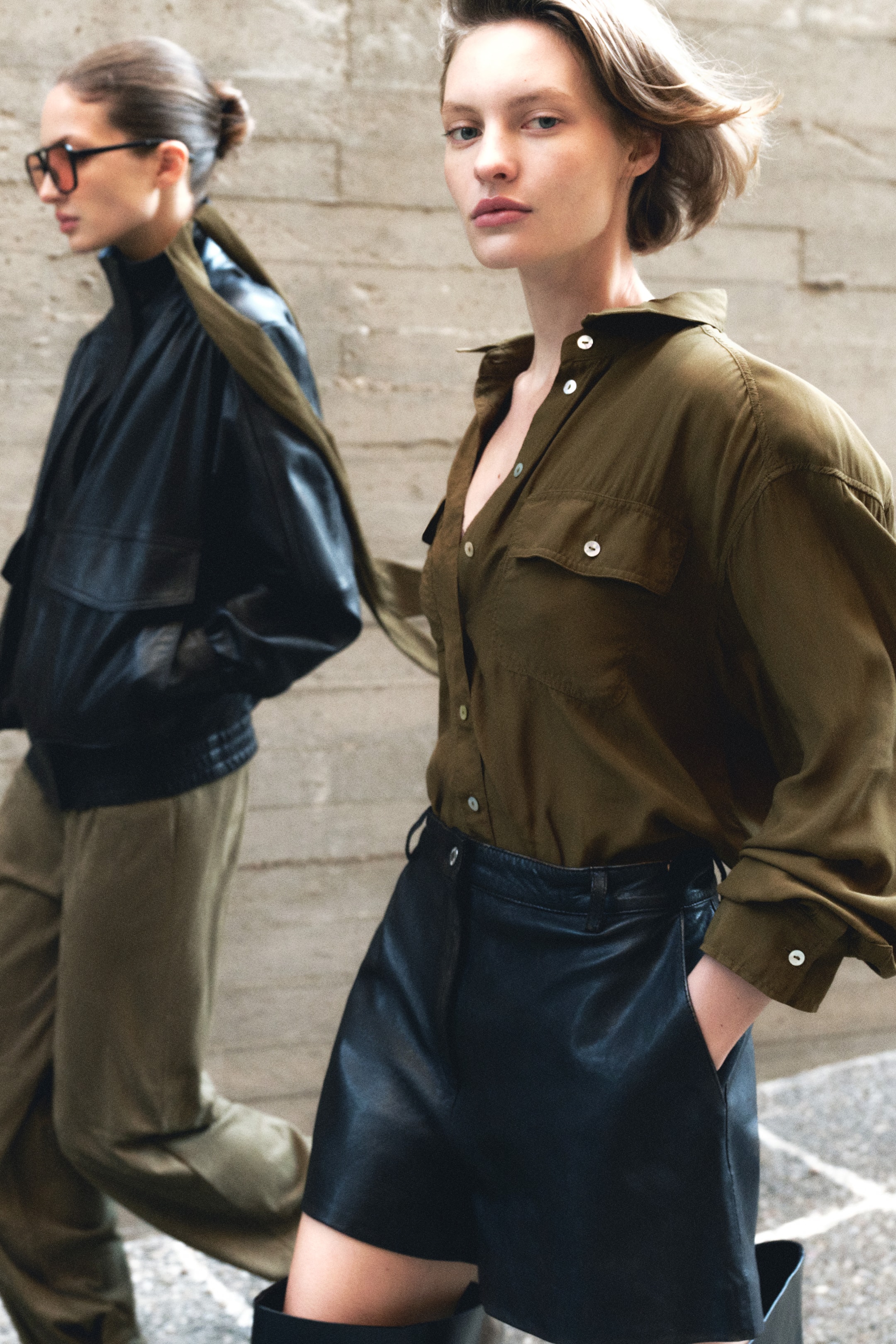 View larger image: A dark olive green button-down shirt features two chest flap pockets and white buttons, with a few buttons left undone at the collar to form a V-neck. The shirt is tucked into high-waisted black leather shorts, which display side pockets and belt loops.