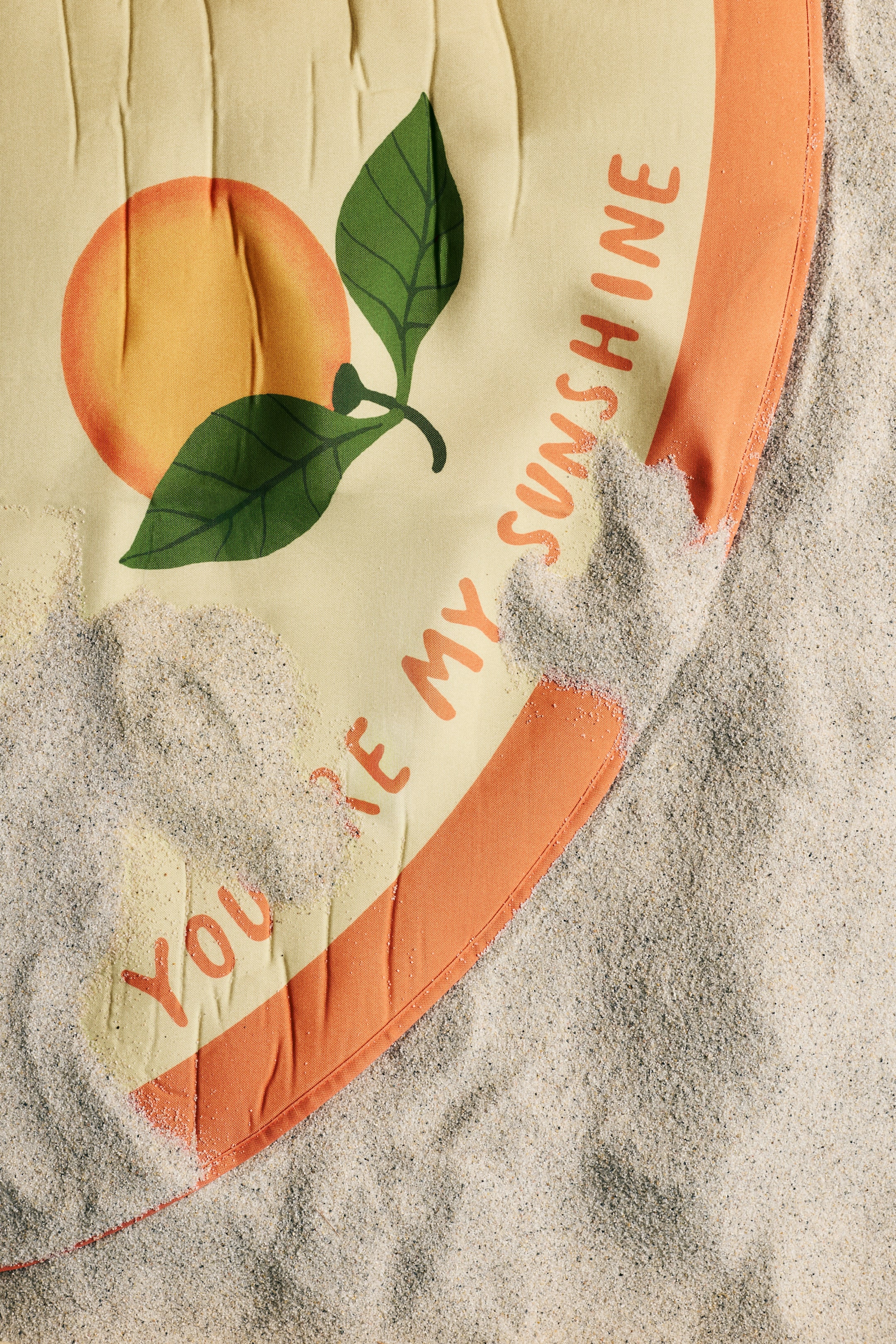 View larger image: This round beach towel showcases a soft yellow background with an orange sun graphic accented by two green leaves. The phrase "YOU'RE MY SUNSHINE" is elegantly curved along the edge in vibrant orange text. The towel's border is also orange, featuring gentle creases, and it is partially nestled in the sand.
