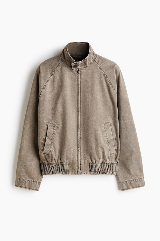 A stone-washed brown jacket is displaying its front, featuring a stand-up collar with a snap button, a central zipper closure, raglan sleeves, two slanted welt pockets, and a ribbed elasticated hem. - 1