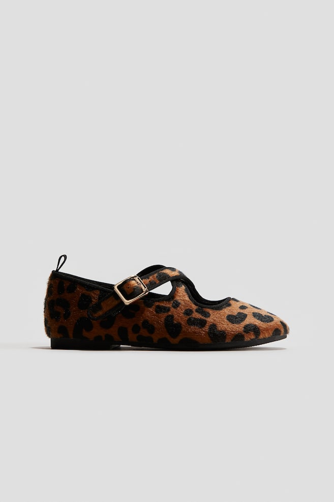 Ein einzelner flacher Mary Jane-Schuh wird in einer Seitenansicht präsentiert und zeigt eine braun-schwarze Leopardenmuster-Ponyhaar-Textur. Er verfügt über einen schwarzen Riemen über dem Spann, der mit einer goldfarbenen rechteckigen Schnalle gesichert ist, eine schwarze Umrandung, die den Schuh und den Riemen umgibt, und eine kleine schwarze Zuglasche, die an der Ferse sichtbar ist. - 1