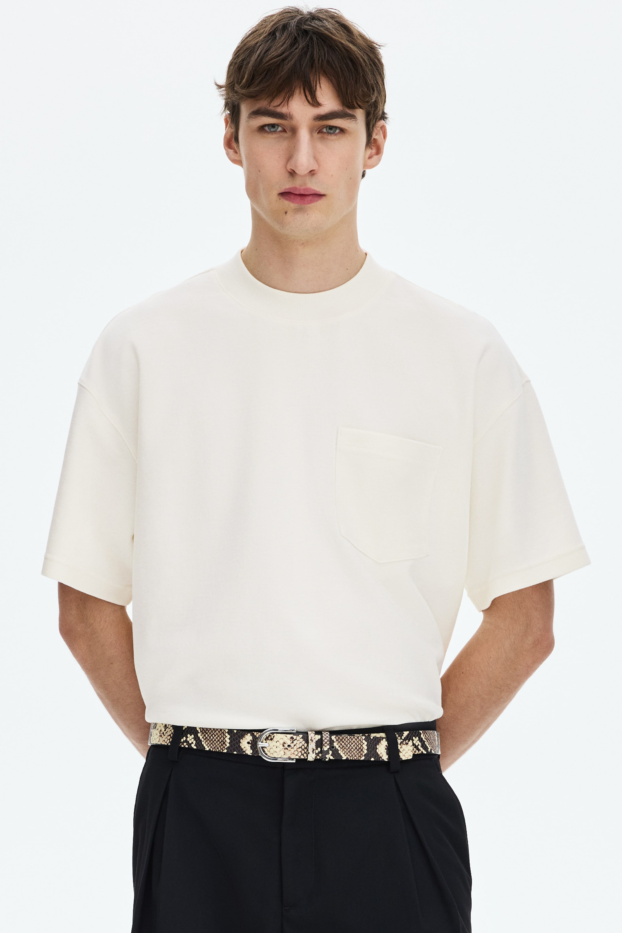 Agrandir l'image: Un t-shirt à manches courtes blanc cassé est présenté de face, doté d'un col montant et d'une coupe décontractée avec des épaules tombantes. Il met en avant une texture en tricot lisse et une seule poche plaquée sur la poitrine gauche.