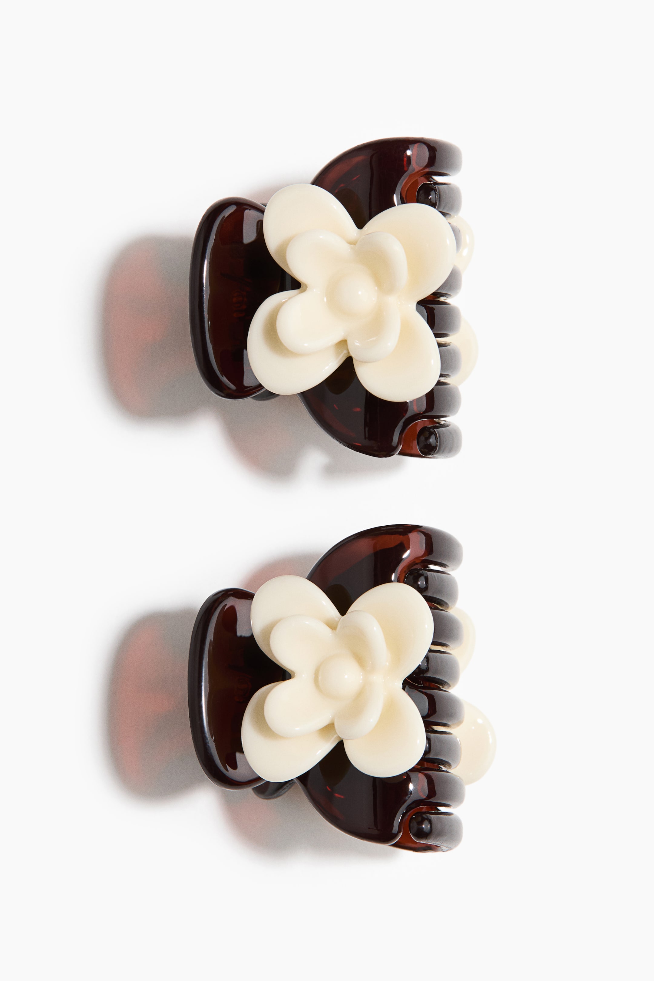 View larger image: Two hair claw clips are arranged vertically, one above the other. Each clip showcases a glossy, dark brown tortoiseshell base with a creamy white, four-petal flower design on its exterior.