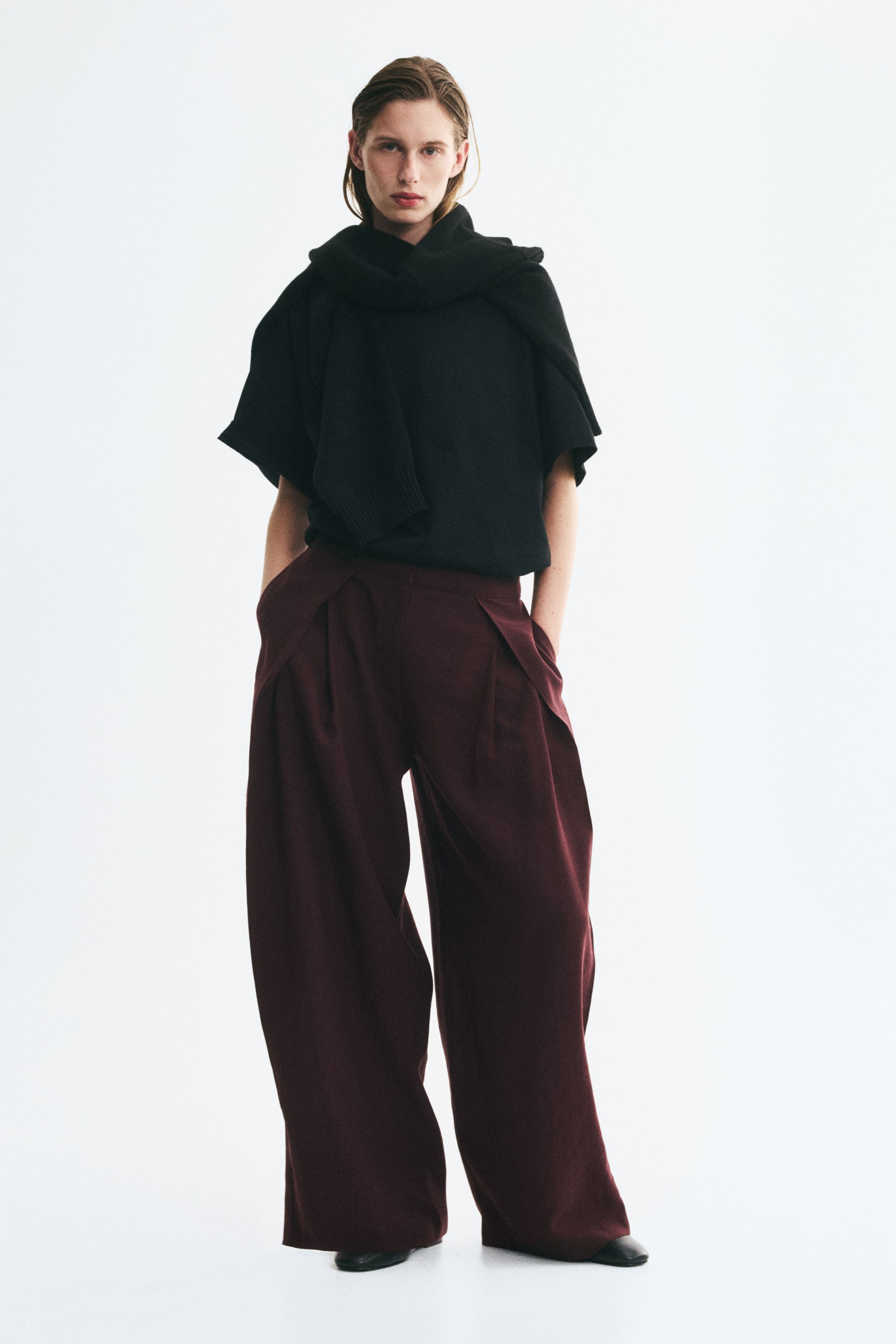 Grotere afbeelding bekijken: Een model draagt een losvallende bordeauxrode broek in een twill van lyocellmix, met de handen in de zijzakken. De broek heeft een wijde barrel-leg-silhouet met plooien vooraan en overlappende zijpanelen, gecombineerd met een zwart topje met korte mouwen en een zwarte gebreide trui nonchalant rond de nek gedrapeerd.