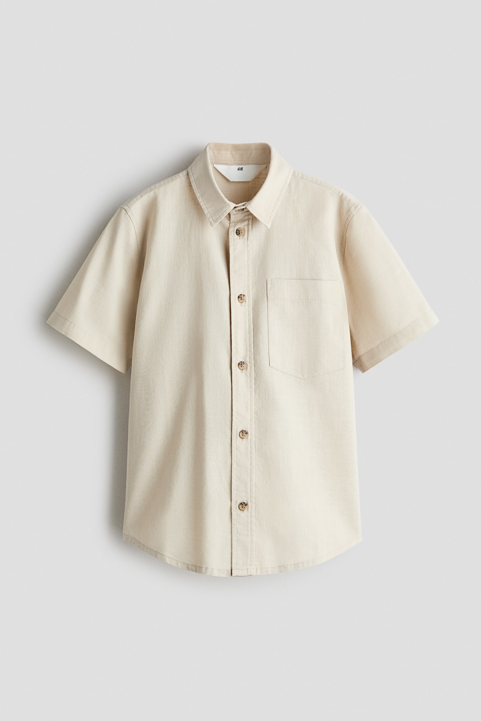 A light beige, short-sleeved button-down shirt is facing front, displaying a textured linen-like fabric, a standard collar, and a single patch pocket on the left chest. It is featuring a vertical row of five brown buttons down the front placket and a slightly curved hem.