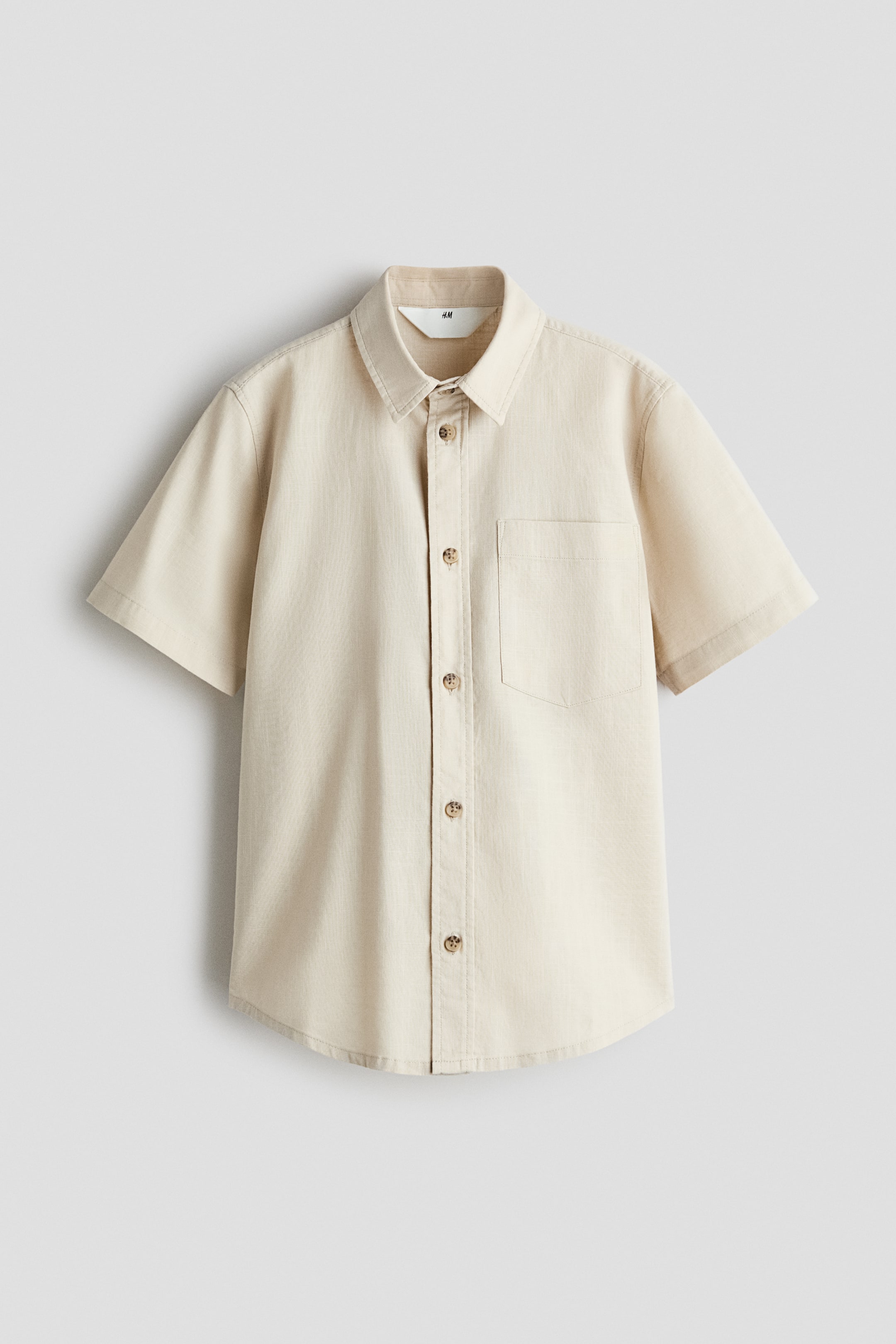 View larger image: A light beige, short-sleeved button-down shirt is shown from the front, featuring a textured linen-like fabric, a standard collar, and a single patch pocket on the left chest. It has a vertical row of five brown buttons down the front placket and a slightly curved hem.