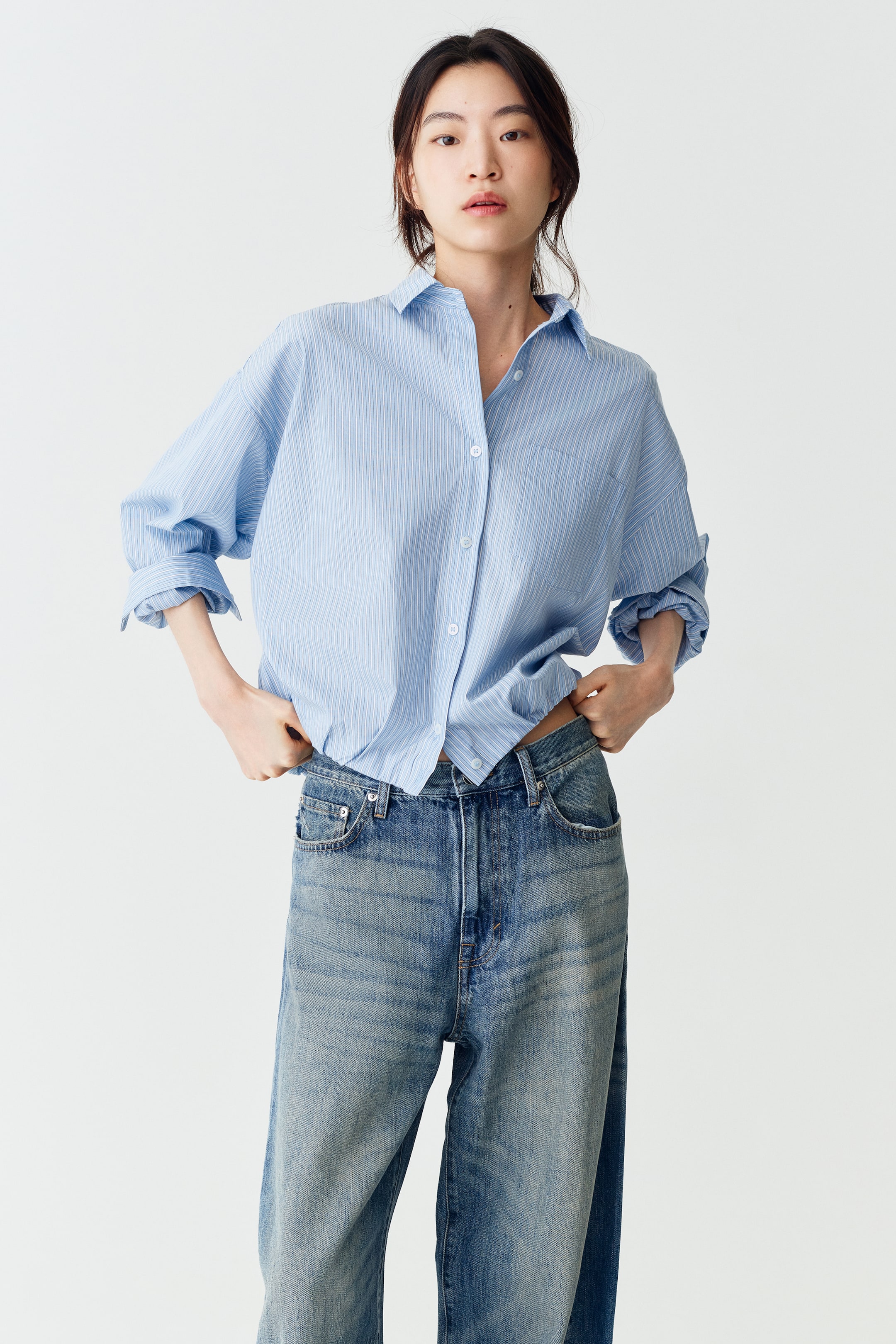 Xem ảnh lớn hơn: Người mẫu đang mặc một chiếc áo sơ mi cotton kẻ sọc xanh nhạt và trắng, có cổ áo, vai rơi và gấu áo phồng kiểu bubble hem. Người mẫu đang cầm hai bên gấu áo tại cạp quần jeans xanh của mình, với tay áo dài của áo được xắn lên đến khuỷu tay.