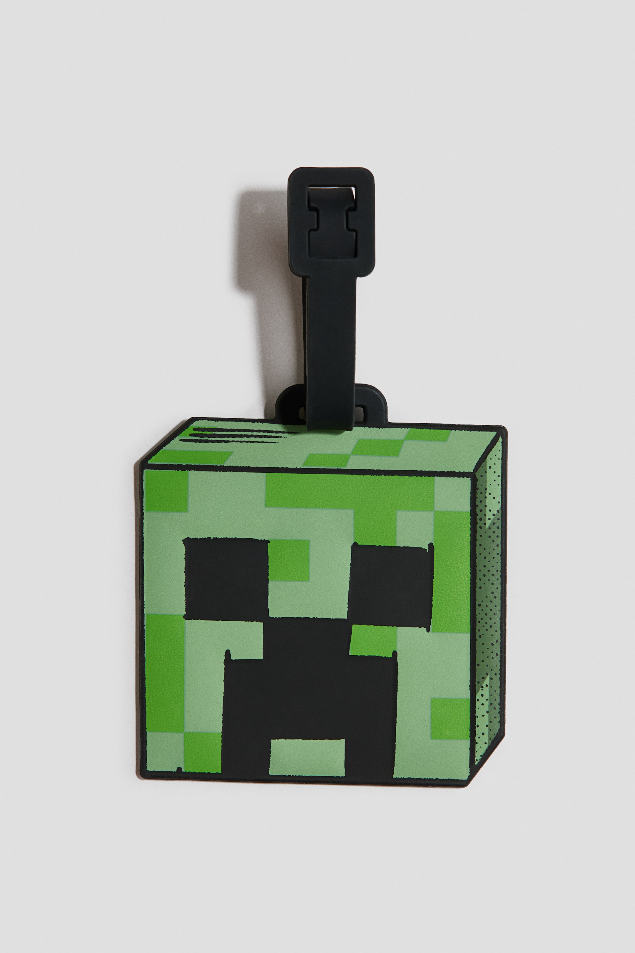 Ver imagem maior: A etiqueta de bagagem apresenta um design frontal de cabeça em bloco, exibindo um padrão pixelizado em vários tons de verde, com pixels pretos formando os olhos e a boca. Uma correia de borracha preta com fivela está presa na borda superior.