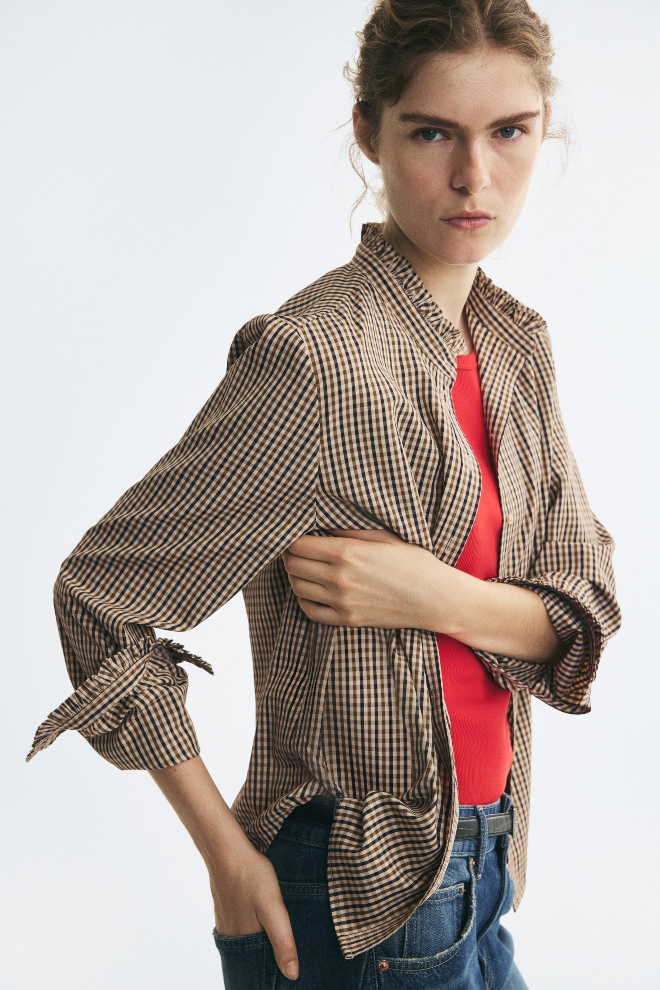 View larger image: A brown and black checkered blouse with a frill-trimmed stand collar is worn unbuttoned over a red top. The model styles the long-sleeved blouse with blue jeans, holding one arm across the chest while resting the other hand in a pocket.