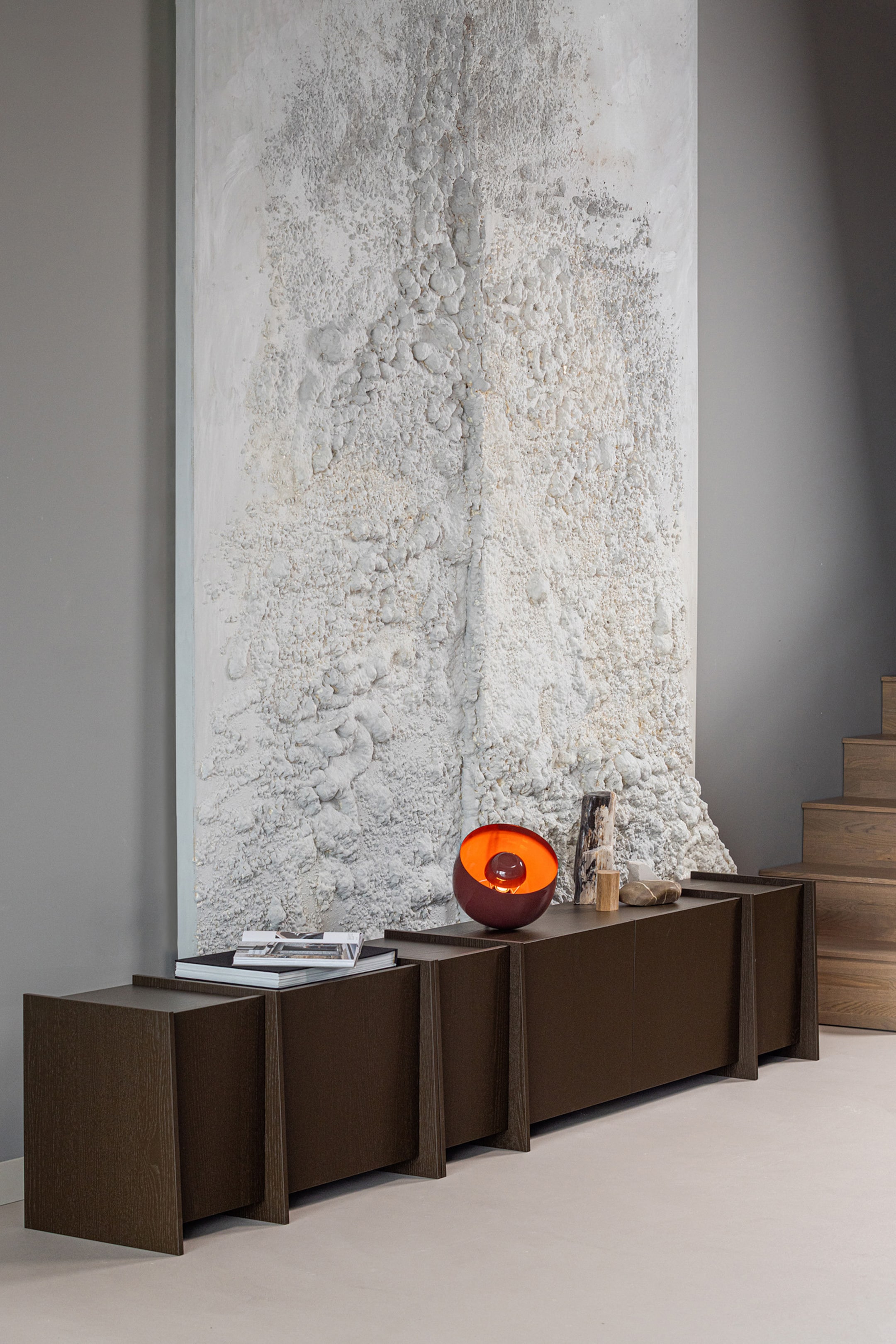 Grotere afbeelding bekijken: Een lange, donkerbruine modulaire kast met meerdere secties en zichtbare houtnerf. Elke sectie rust op schuine, trapeziumvormige zijkanten en het vlakke bovenoppervlak toont boeken, een lamp en decoratieve objecten.