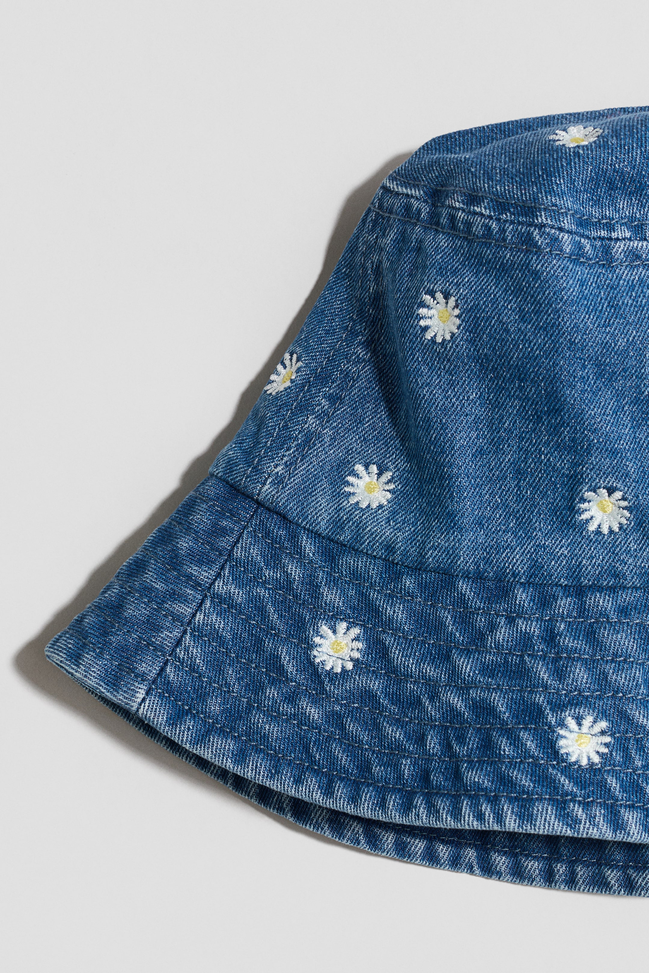 Visualizza l'immagine ingrandita: Un cappello a secchiello in denim blu è caratterizzato da piccoli ricami di margherite bianche con centri gialli. Il suo ampio bordo presenta più file di cuciture concentriche.