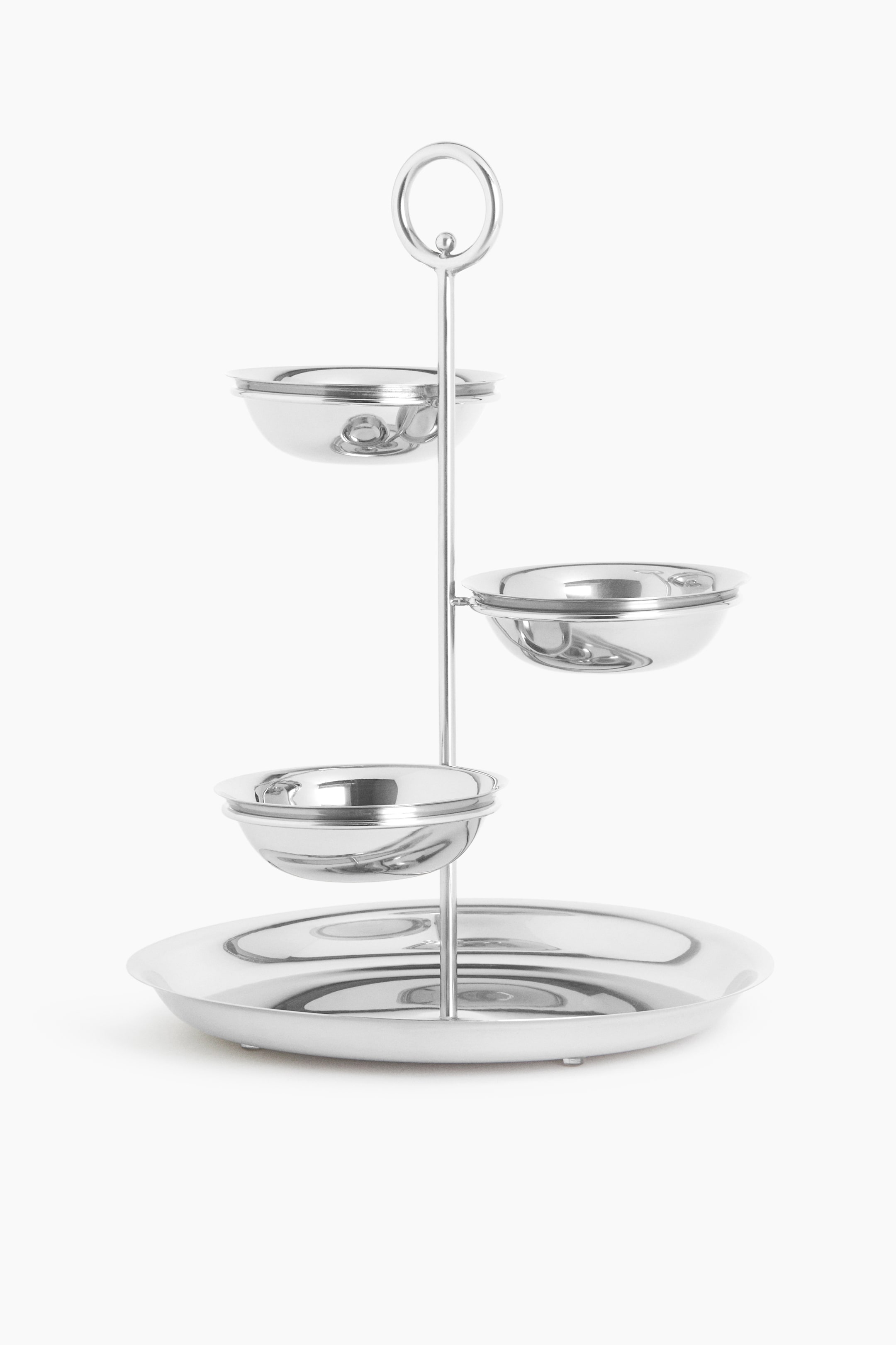 View larger image: A polished silver-toned metal tiered stand is positioned facing front, featuring a large circular tray at the base and a central vertical rod with a ring-shaped handle at the top. Three small, matching bowls extend outwards from the central rod at staggered heights in a spiralling arrangement.