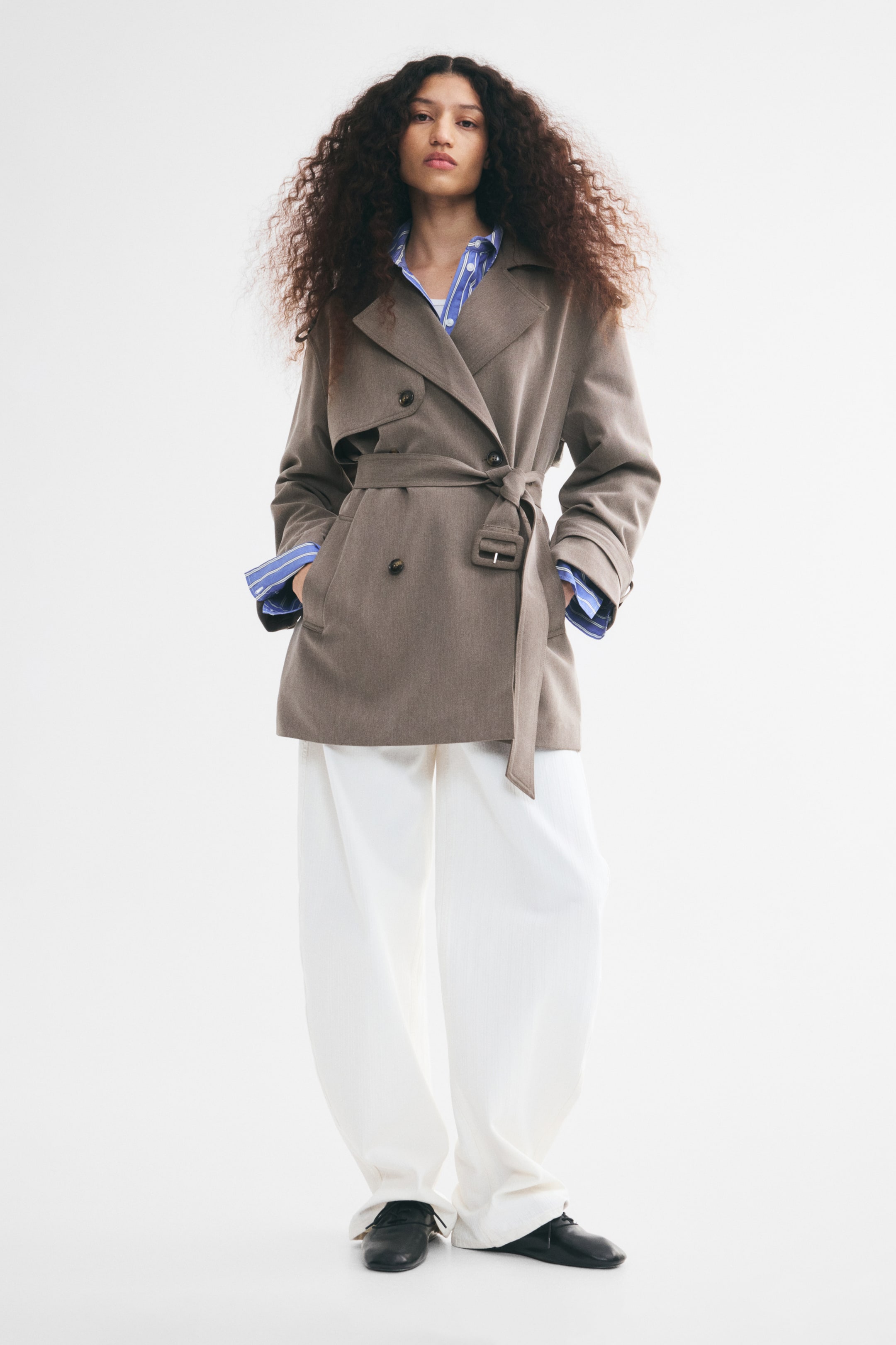 View larger image: A short, double-breasted trench coat in taupe twill is being worn by the model with their hands placed in the front welt pockets. The oversized-fit coat features notch lapels, a storm flap, and an adjustable waist belt tied at the front, and is styled over a blue and white striped collared shirt and wide-leg white trousers.