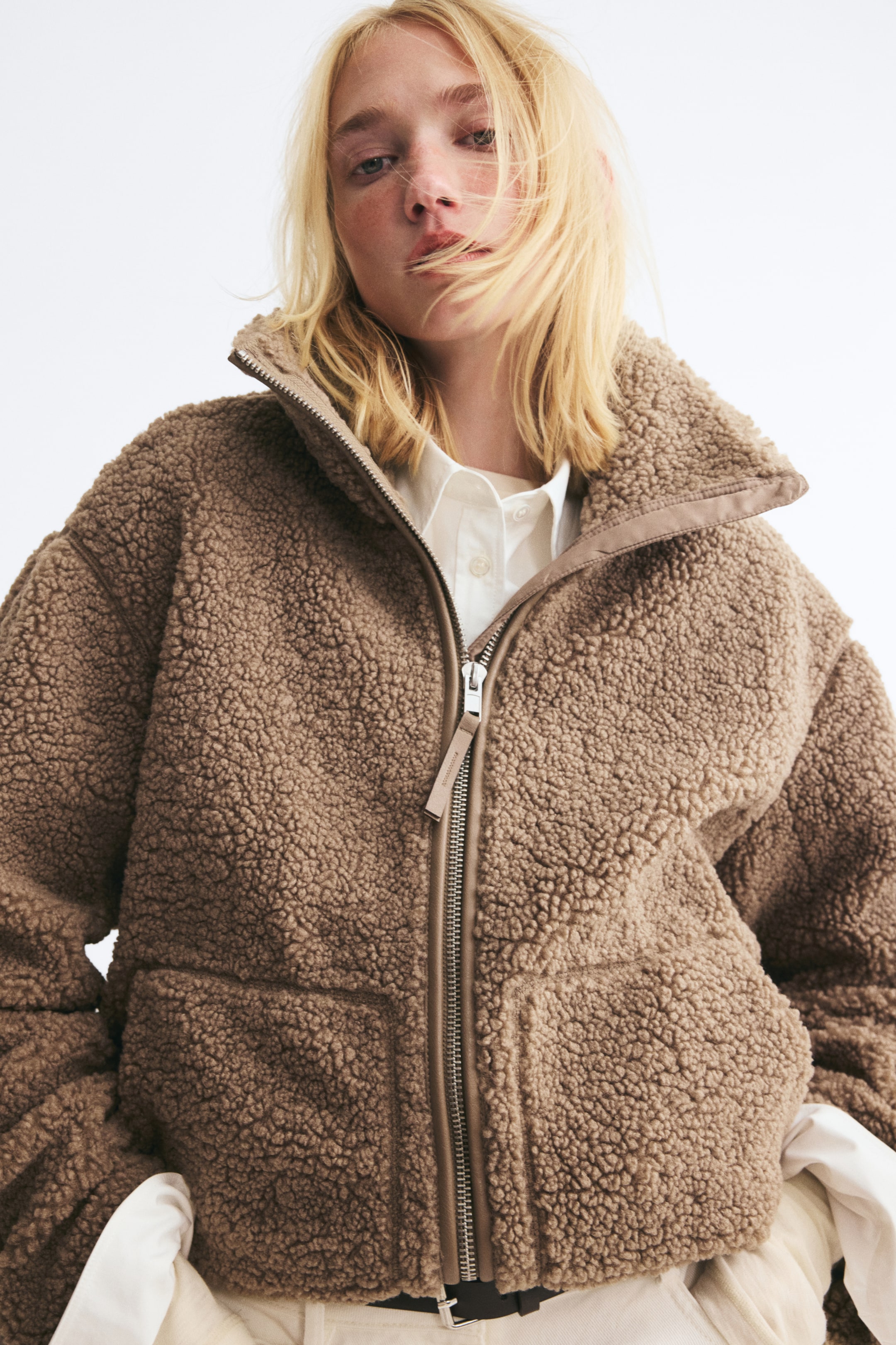 View larger image: An oversized brown teddy jacket is being worn by the model, layered over a white button-down shirt. The jacket features a funnel collar, a front zip closure with coated fabric trim, and large side pockets.