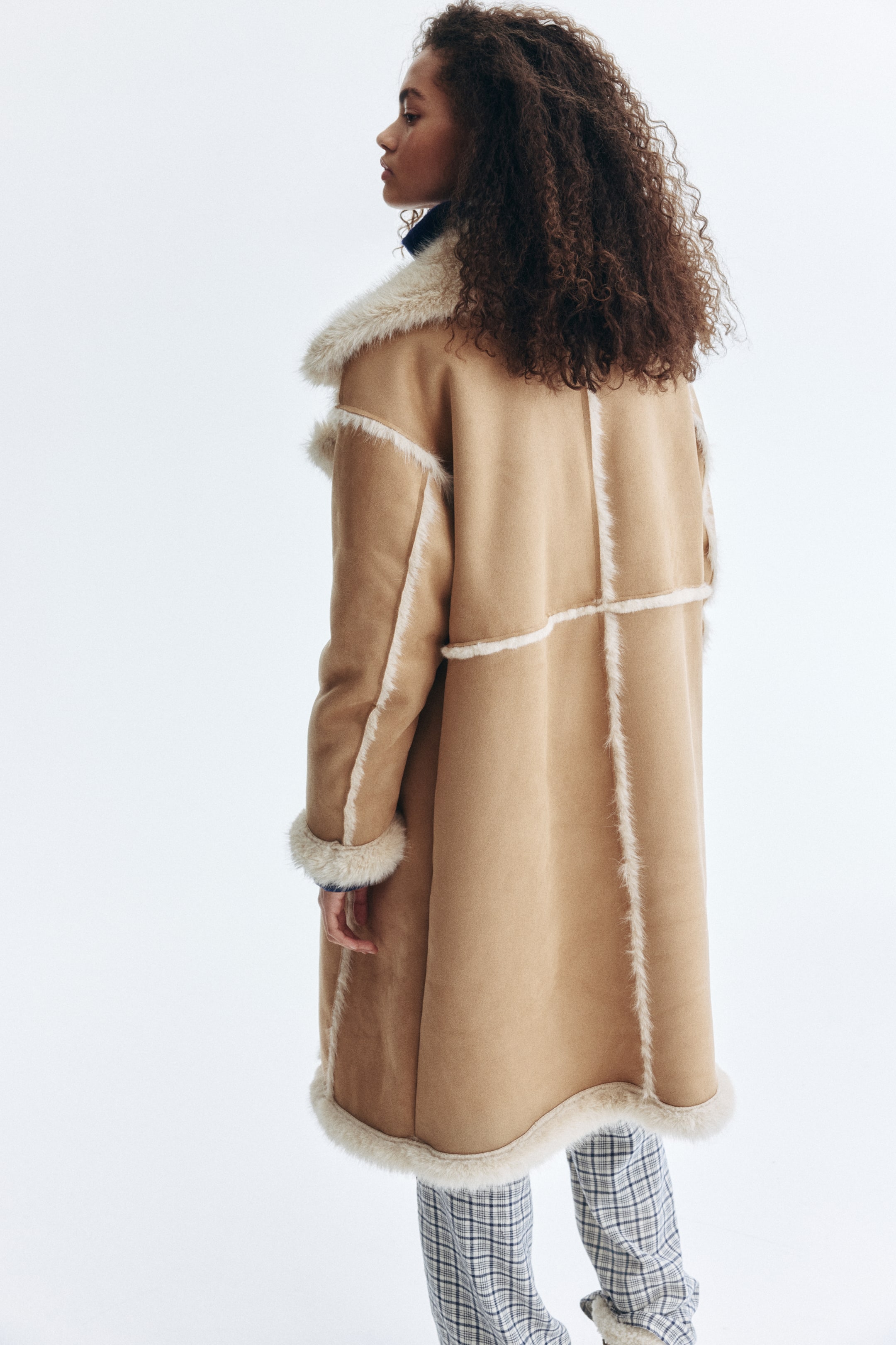 Agrandir l'image: Un long manteau brun clair avec une finition douce et moelleuse est porté par le mannequin. Il présente des garnitures duveteuses de couleur claire le long des coutures, des poignets et de l'ourlet, ainsi qu'un col duveteux, révélant sa doublure intérieure duveteuse.