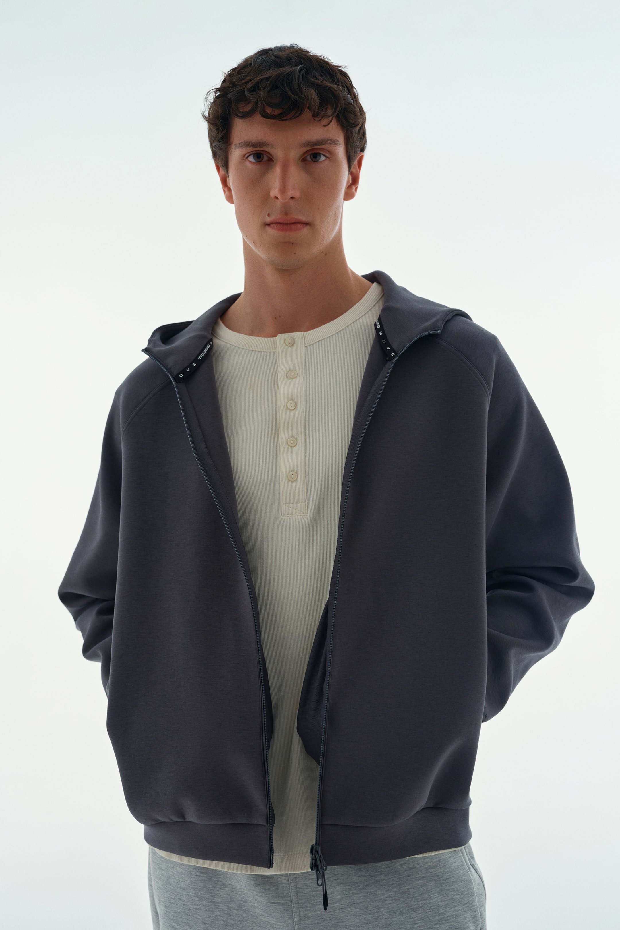 Grotere afbeelding bekijken: Een donkergrijze hoodie met ritssluiting wordt open gedragen door het model, dat de handen in de steekzakken heeft. De hoodie heeft raglanmouwen en is gelaagd over een gebroken wit henley-overhemd met knoopsluiting.