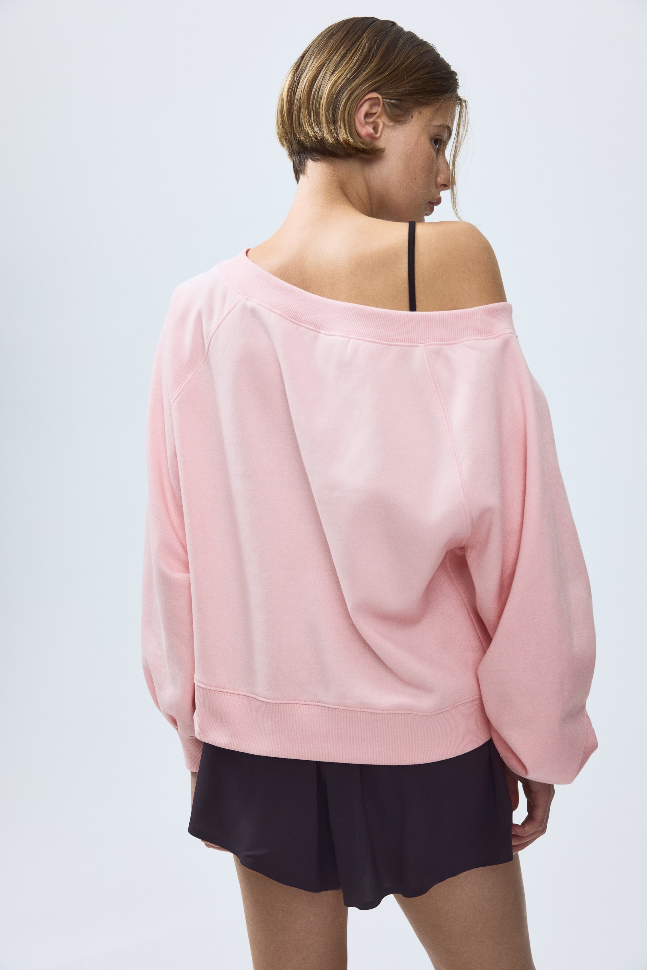 View larger image: The model is wearing a light pink, loose-fit off-the-shoulder top made from sweatshirt fabric, featuring long balloon sleeves with a raglan cut and ribbed cuffs and hem. The top's wide neckline is pulled down, revealing the model's left shoulder.