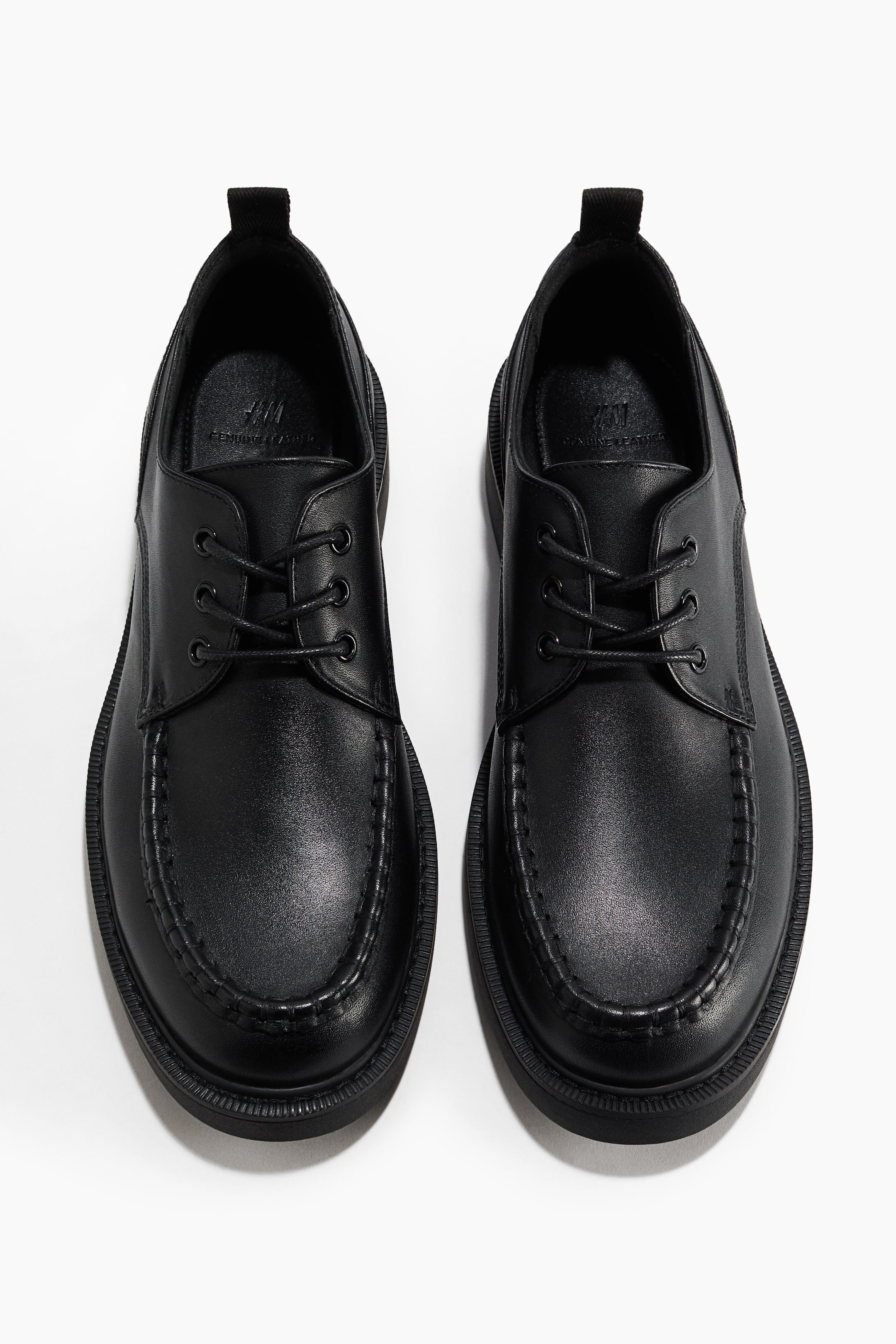 View larger image: A pair of black leather moc-toe derby shoes is shown front-facing in a top-down view, featuring a three-eyelet lace-up design with matching black laces and prominent whipstitching around the toe. The shoes have thick, ridged black rubber soles and fabric pull tabs at the heels.