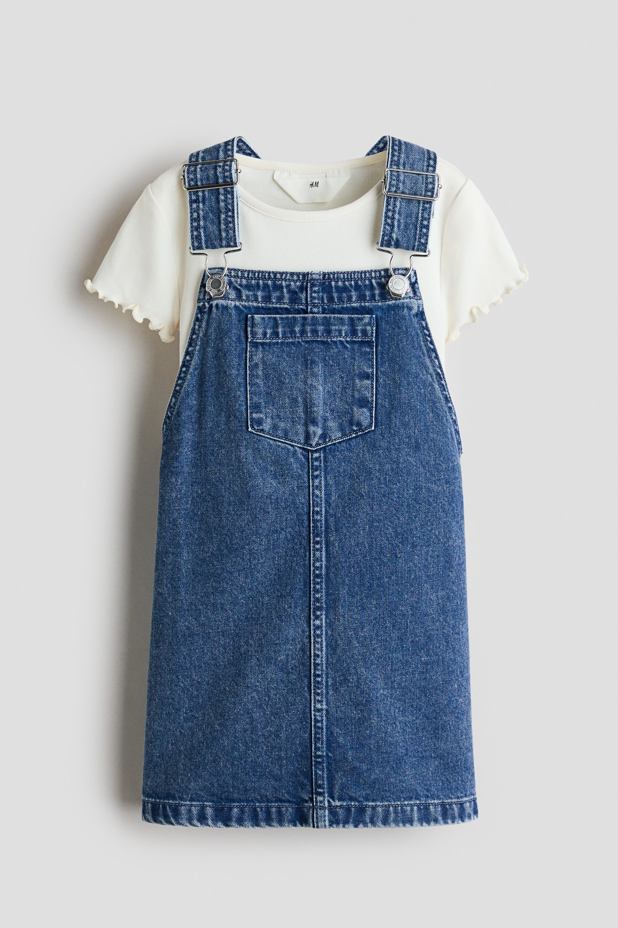 View larger image: A blue denim overall dress is shown front-facing, featuring adjustable shoulder straps with silver-toned metal buckles and a front bib with a patch pocket. It is layered over a cream short-sleeved T-shirt with a round neck and lettuce-edge-trimmed sleeves.