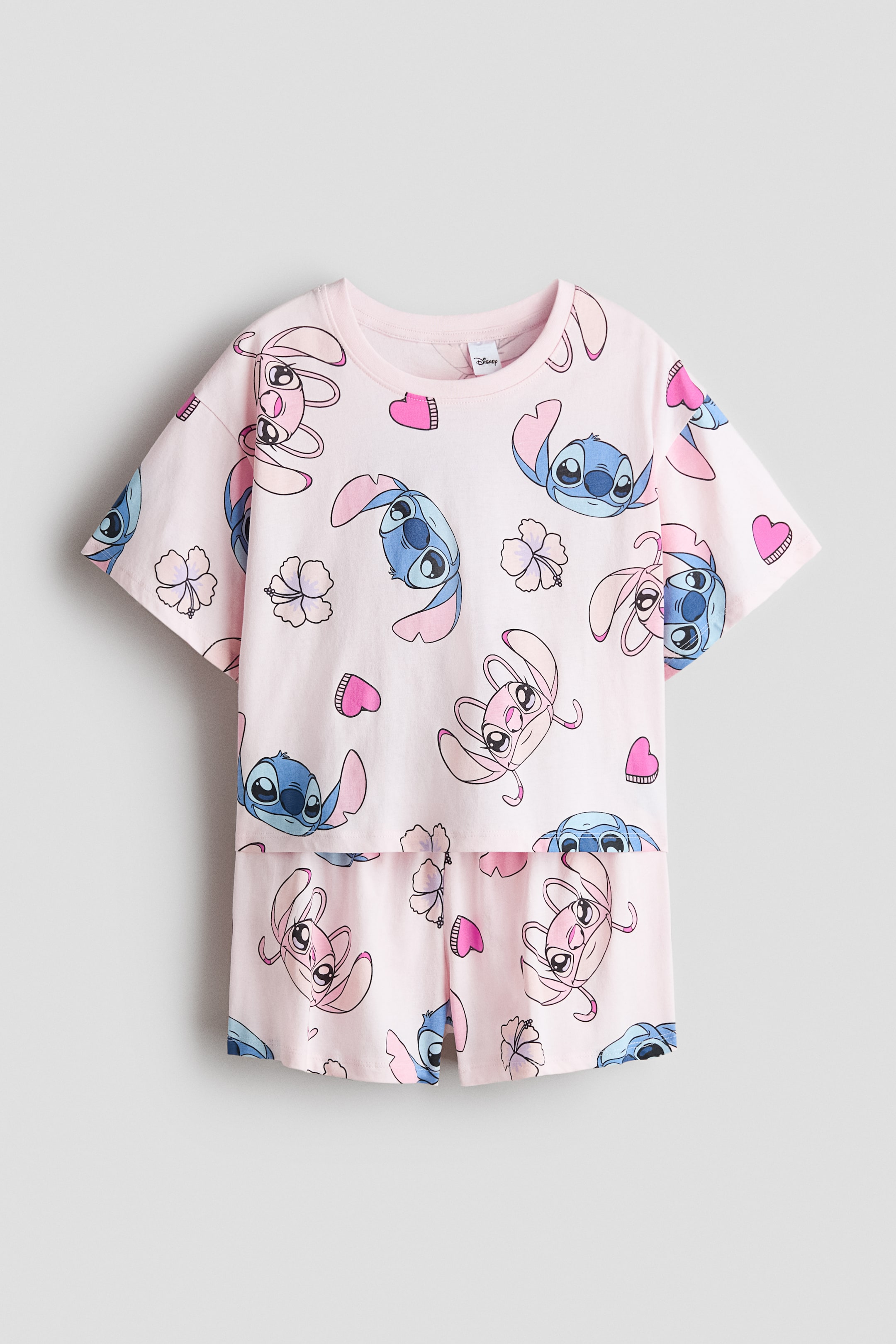 View larger image: Pink cotton jersey pyjamas are laid out flat, front facing, showcasing an all-over print of Stitch and Angel characters, pink hearts, and light flowers. The T-shirt features a ribbed neckline and dropped shoulders, paired with matching shorts.