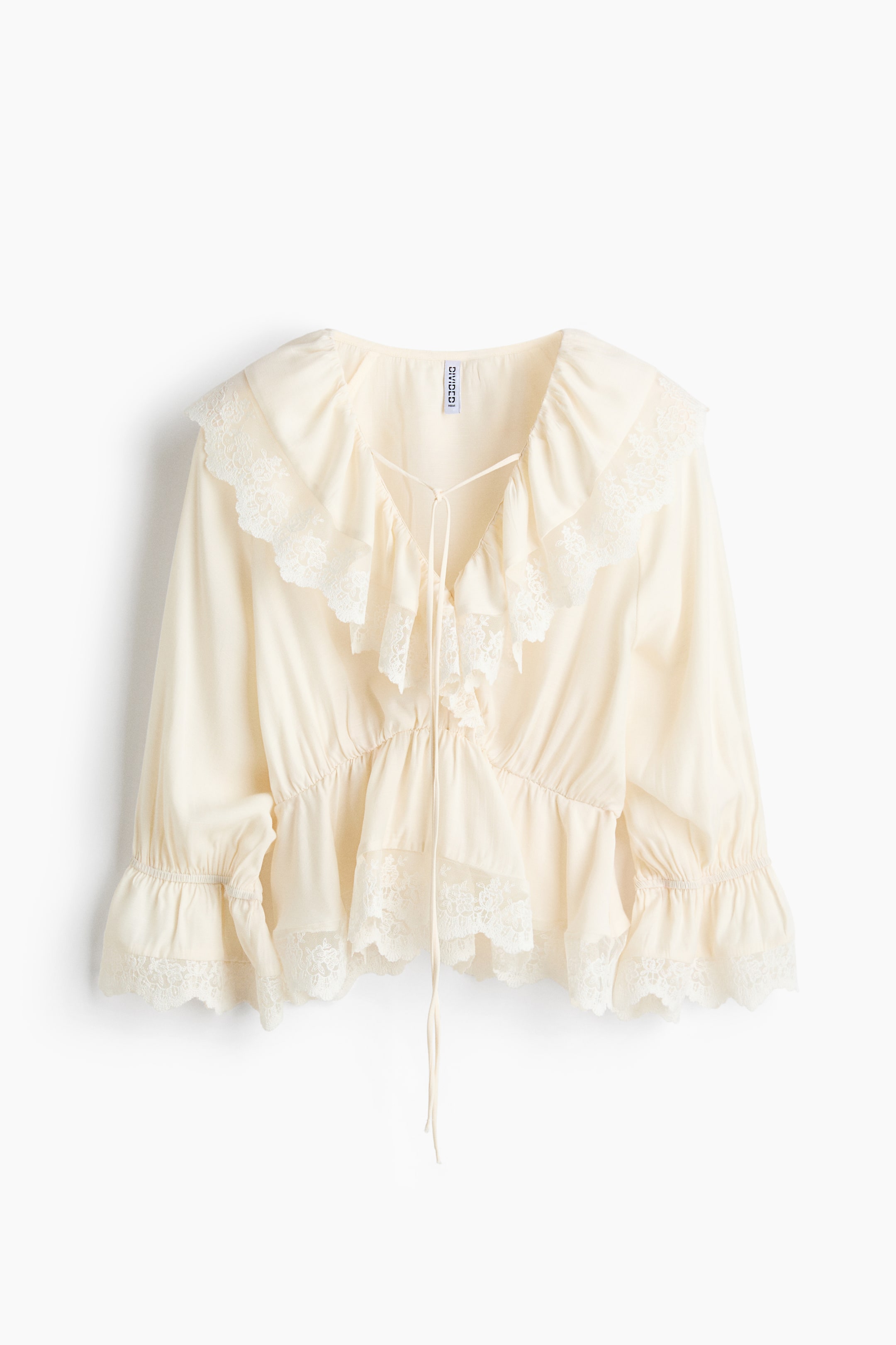 View larger image: A cream-colored blouse features a wide ruffled collar with lace trim, framing a V-neckline with ties. The blouse showcases long sleeves with gathered, lace-trimmed cuffs and a gathered peplum waistline with matching lace detailing.