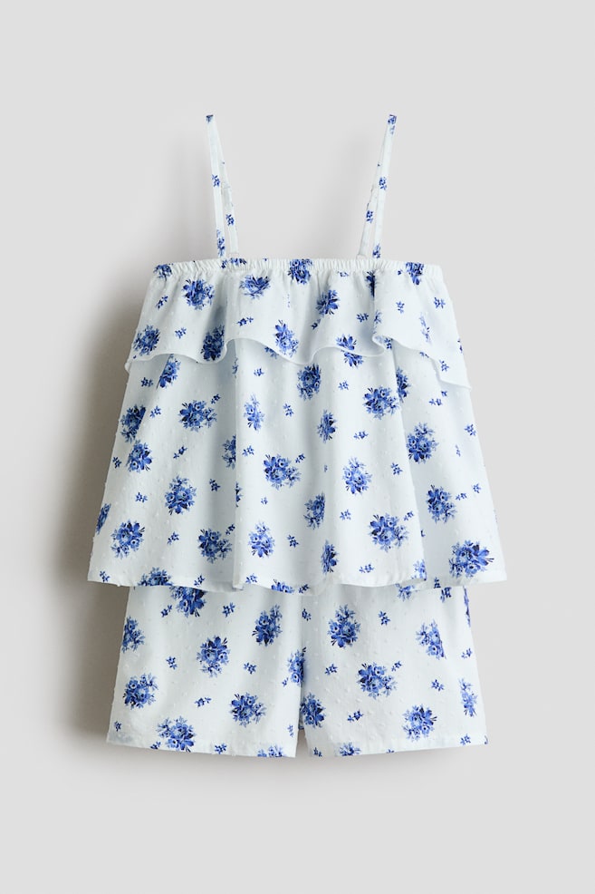 A white two-piece set, comprising a top and matching shorts, is displaying a blue floral print and small raised dots. The top is featuring spaghetti straps, an elasticized neckline, and tiered ruffles. - 1