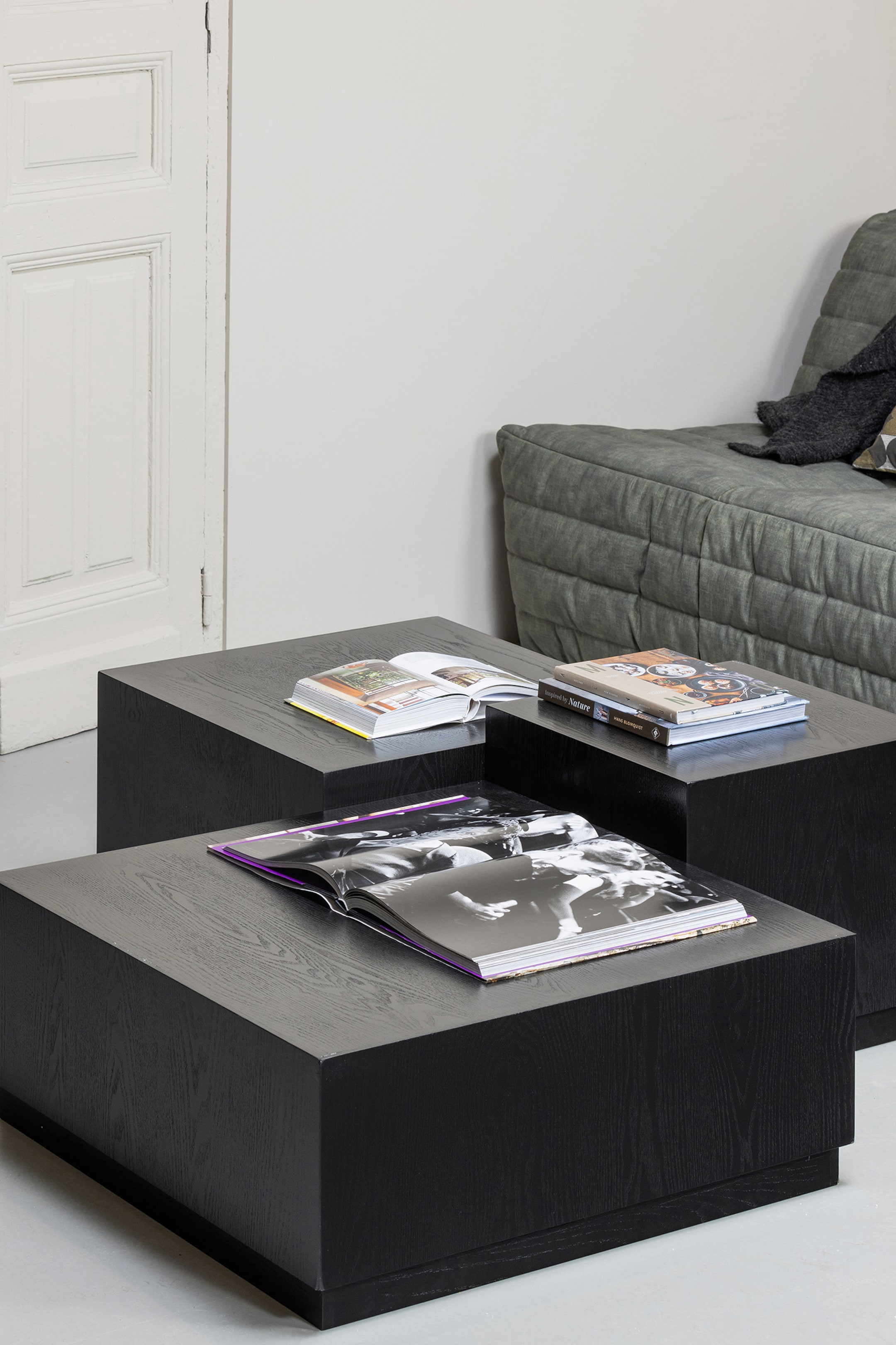 Grotere afbeelding bekijken: De zwarte, rechthoekige salontafels tonen een zichtbare houtnerfstructuur, waarbij één tafel lager en voor een hogere tafel is geplaatst. Open boeken liggen op hun bovenoppervlakken.