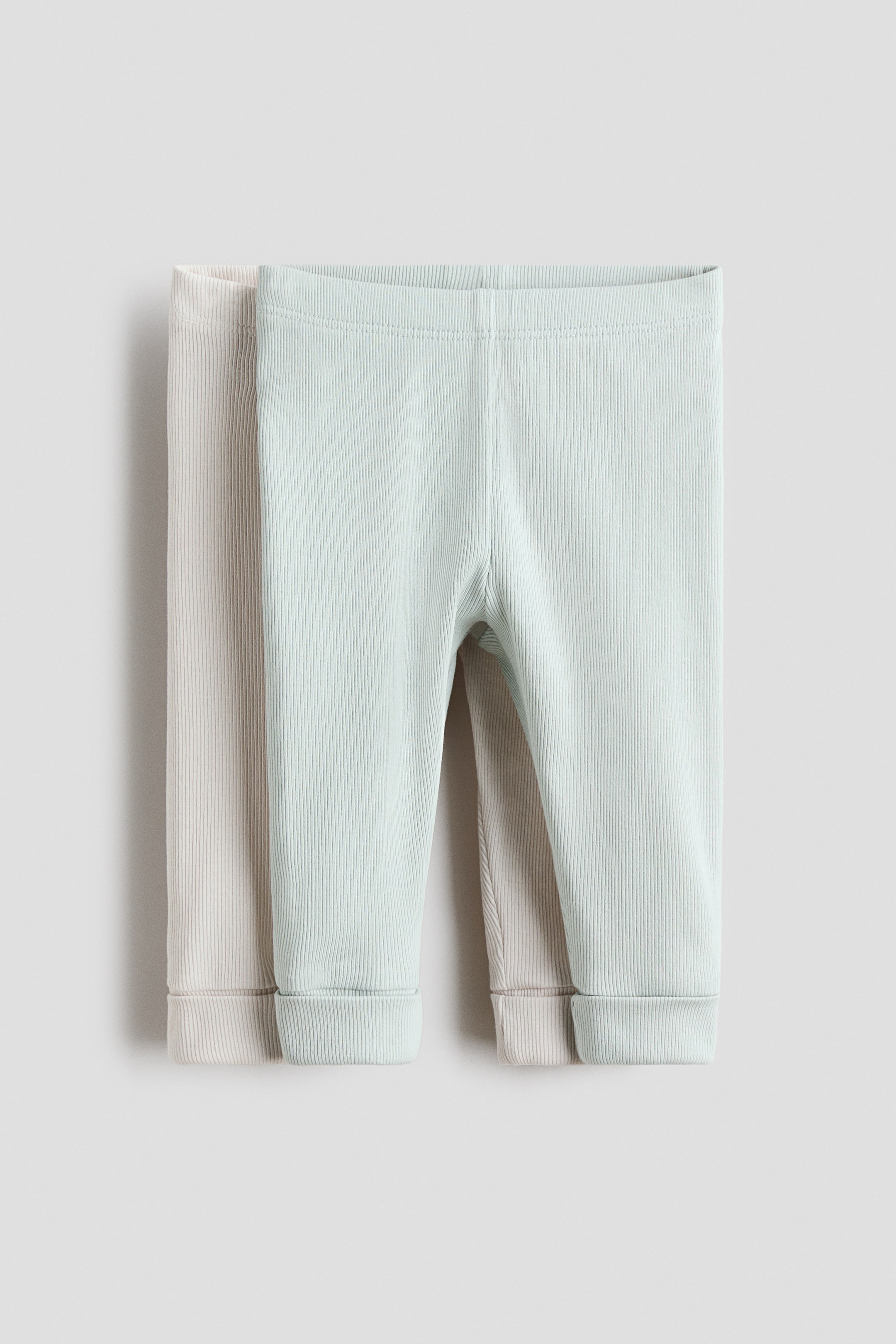 Visualizza immagine più grande: Due paia di leggings a coste sono mostrati dal davanti, con un paio verde chiaro stratificato su un paio beige chiaro. Entrambi i leggings presentano cinture lisce e orli con risvolto.