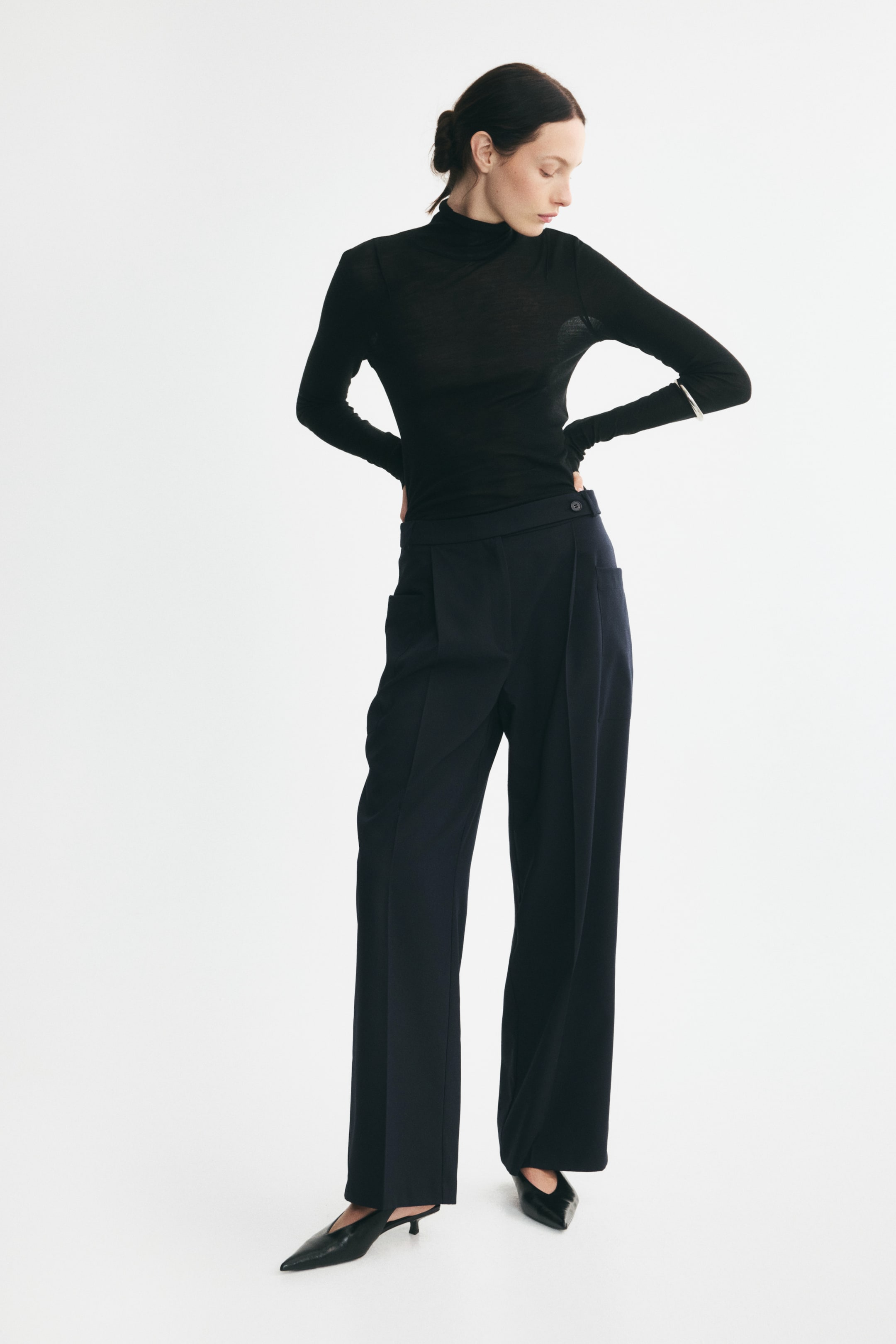 Zobacz większy obraz: Luźne spodnie o szerokich nogawkach z ciemnej tkaniny, noszone przez modelkę, z zaprasowanymi kantami z przodu oraz wydłużonym pasem zapinanym na guzik z boku. Spodnie zestawiono z włożonym w środek czarnym golfem z długim rękawem oraz czarnymi szpilkami z noskiem w szpic.