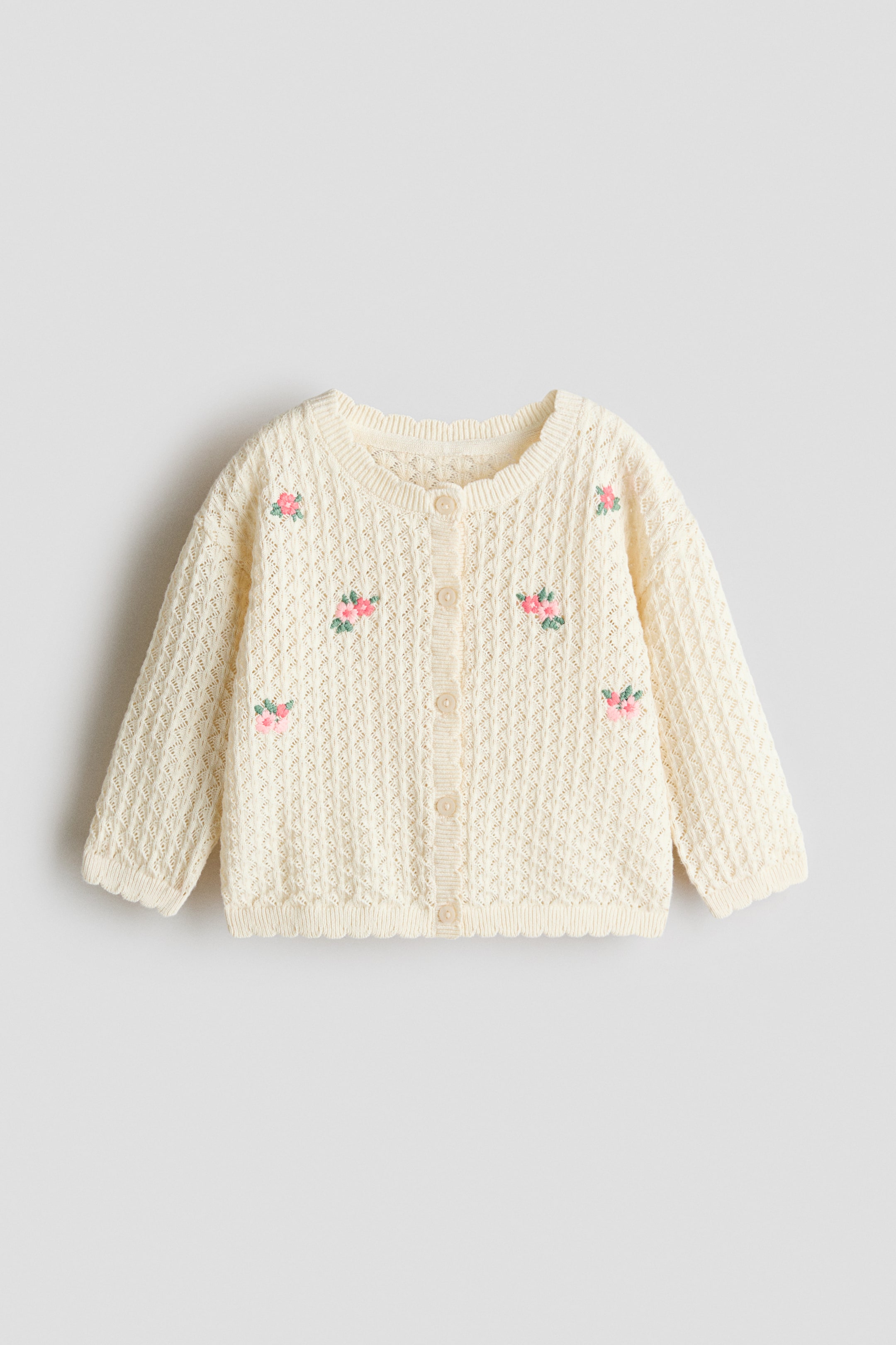 View larger image: A cream baby cardigan is shown from the front, featuring an openwork knit pattern adorned with embroidered pink and green floral clusters, a round neckline, button-through front fastening, ribbed cuffs, and a scalloped hem.