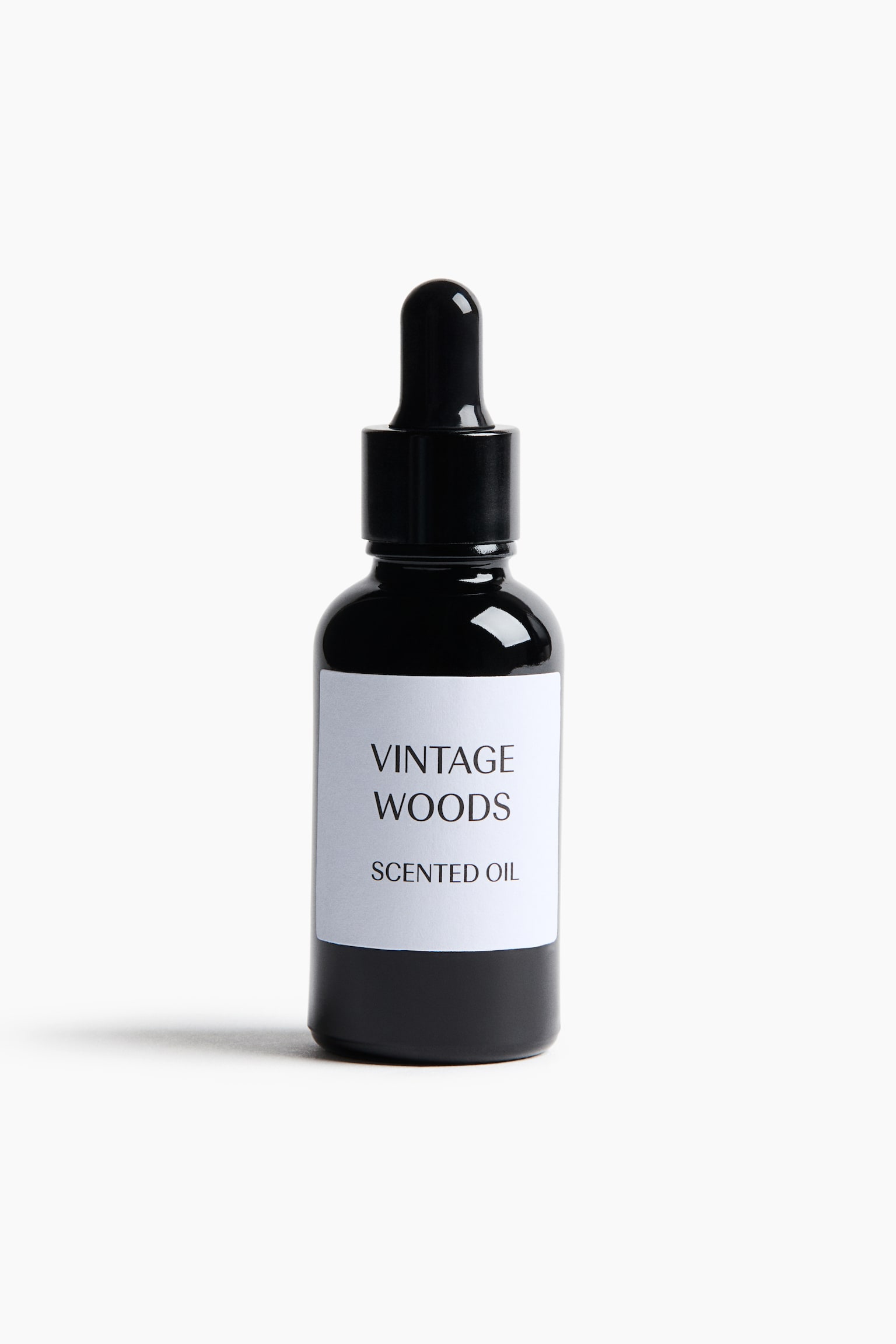 A bottle of scented oil stands upright, facing forward, showcasing a dark, glossy black glass body with a matching black dropper cap. A white rectangular label is affixed to the front, displaying the text "VINTAGE WOODS" and "SCENTED OIL" in a clean black serif font.