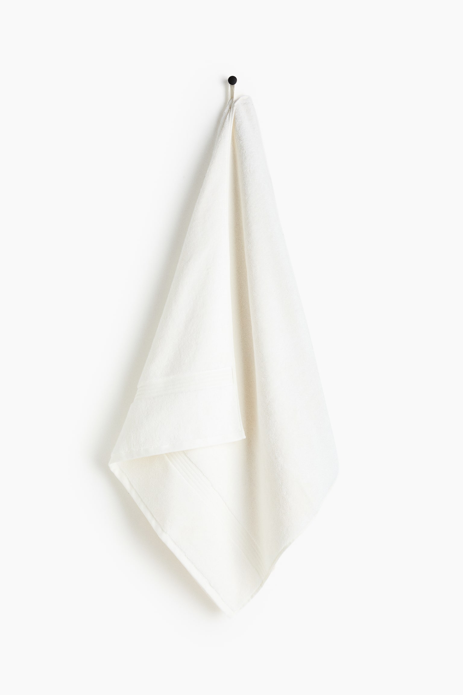 An off-white towel is folded in half lengthwise, draping over a black hook and displaying subtle textured bands near its bottom edge. - 1