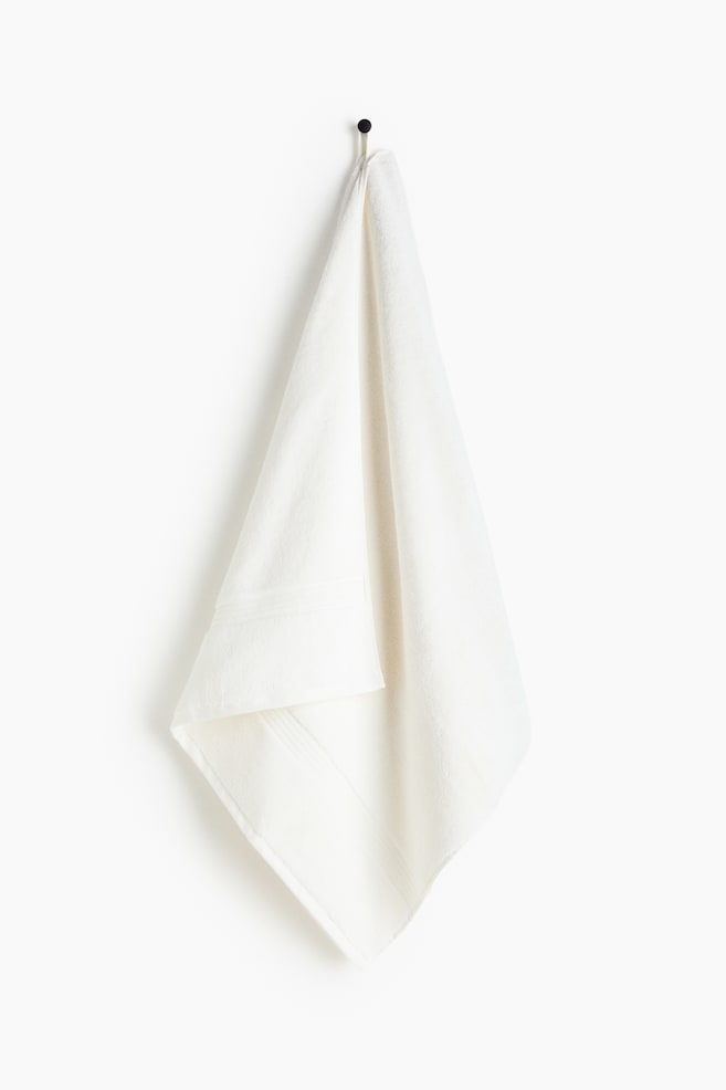 An off-white towel is folded in half lengthwise, draping over a black hook and displaying subtle textured bands near its bottom edge. - 1