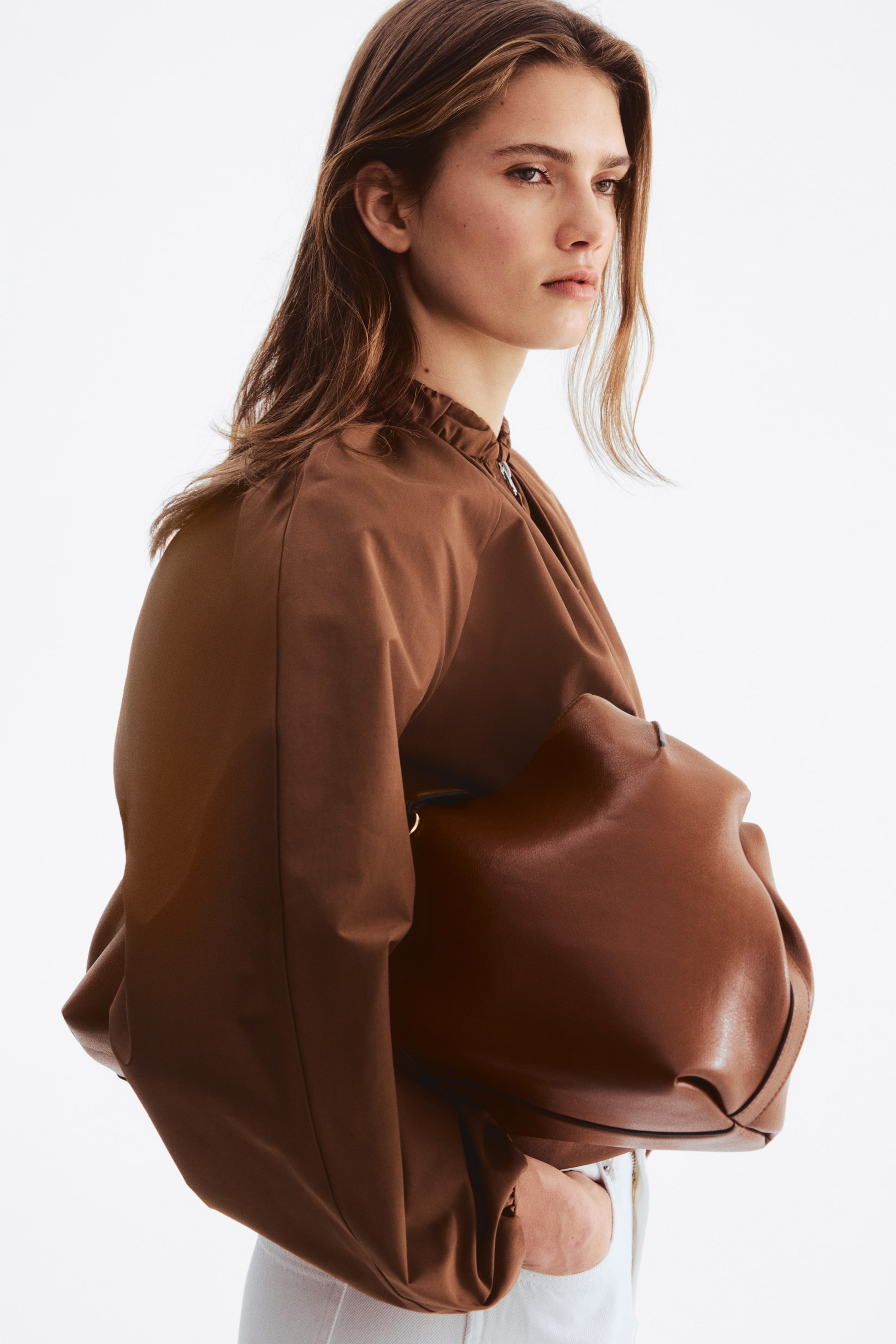 View larger image: A soft brown leather bag is being held against a brown top, showcasing its supple material and slouchy, rounded shape. It features a smooth finish and a subtle gold-toned ring detail near the top opening, emphasizing its flexible structure.